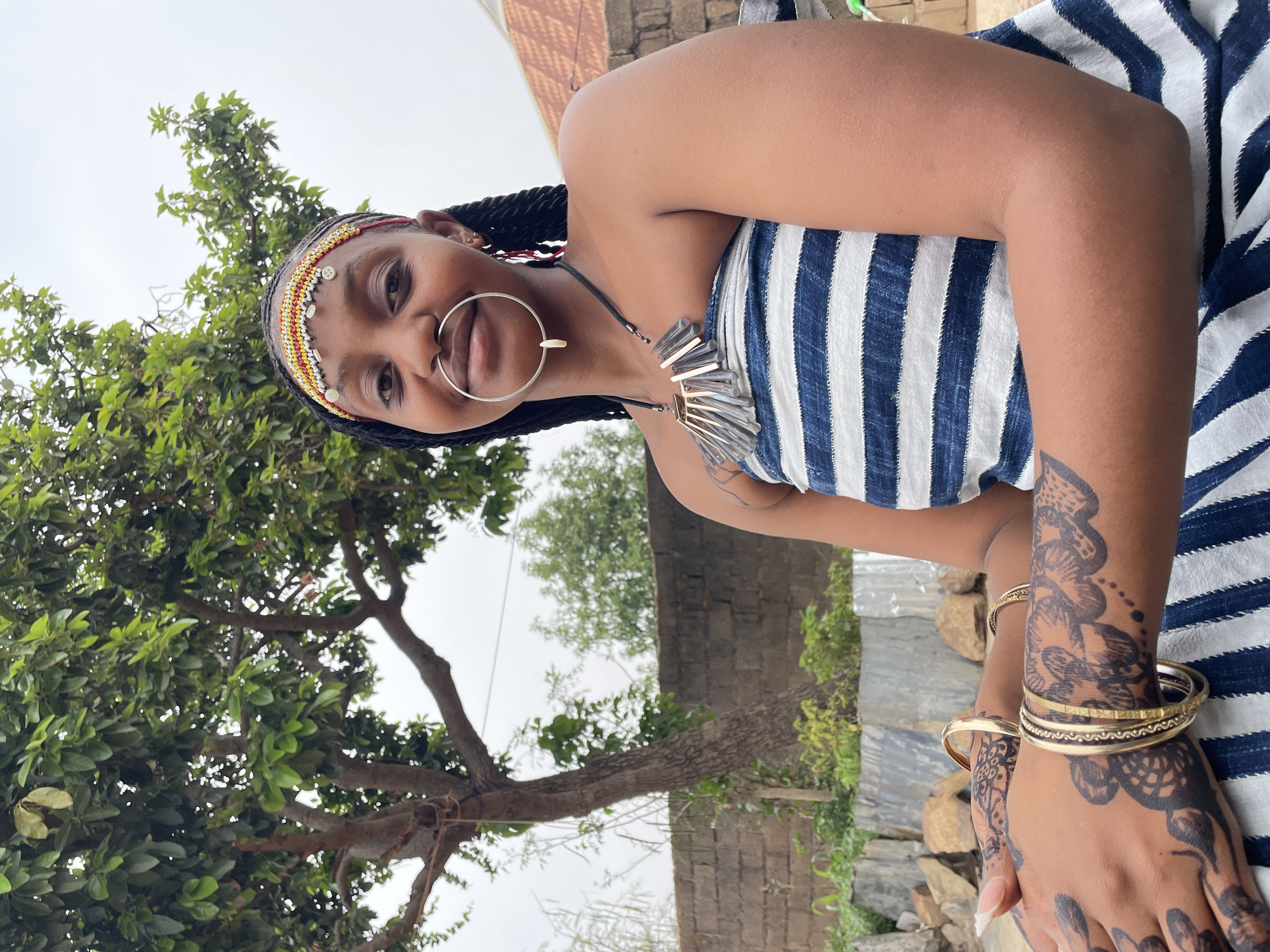 A medium shot depicts a woman outside in what appears to be a yard. The woman, positioned off-center, is adorned in traditional jewelry, including a large nose ring, a beaded headband, and a statement necklace. She is wearing a strapless top with blue and white stripes. Her arms are embellished with henna designs and gold bangles. To the left of the woman, a tree with green leaves partially obscures the background. Behind the tree, a wall constructed of stone is visible. The sky is overcast, casting soft light over the scene. The location is Jos, Nigeria.