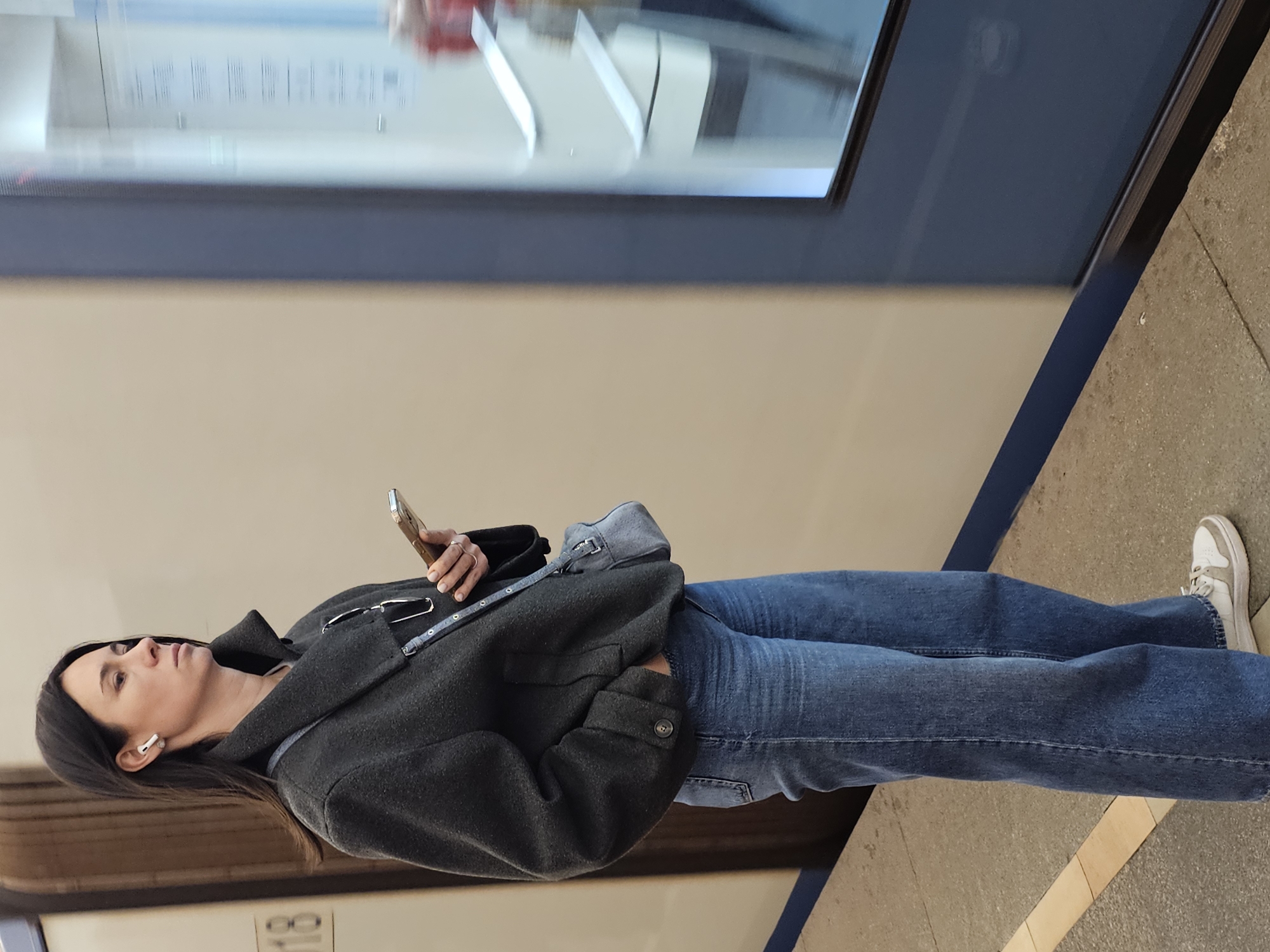 A woman is standing in what appears to be a subway station. She is wearing a dark gray coat, blue jeans, and white sneakers. She has dark hair and is wearing a white earbud in her left ear. She is holding a phone in her right hand and looking up at something out of frame. The background includes a train with a blue stripe and a window, as well as a tiled floor. The location is listed as Unknown, Unknown.