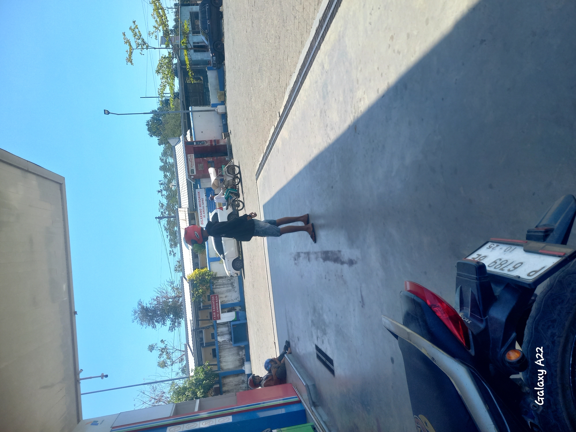 The image captures a scene at a gas station in Kotakan, Indonesia, under a clear blue sky. In the foreground, the rear of a motorcycle is visible, displaying a license plate that reads "P 6789 BE 10-25". To the left, a blue and red gas pump structure stands. Further into the scene, two individuals are present. One person, wearing a red helmet, is seated near the gas pump, holding another helmet. Another individual, also wearing a red helmet and a dark top with denim shorts, stands to the right, facing away from the camera. In the background, a white car is parked, and a person is riding a three-wheeled motorcycle-like vehicle. Beyond the vehicles, buildings with a white facade and blue trim are visible, surrounded by trees. A street lamp stands prominently in the middle ground. The ground is paved with concrete, marked with faint stains and a metal grate running across it.