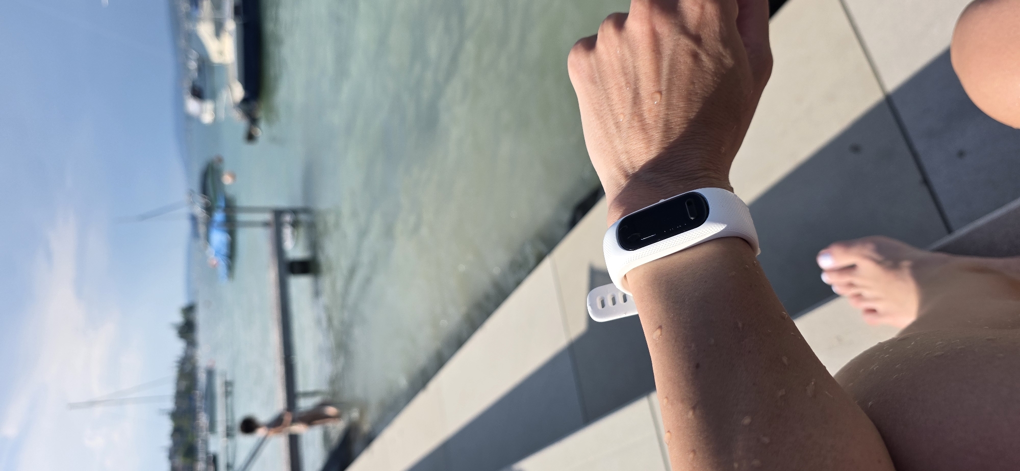 The image is a close-up shot of a person's arm wearing a white fitness tracker, with their feet visible in the foreground. The scene appears to be outdoors, next to a body of water. In the background, a person is standing on a dock, and several boats are visible on the water. The sky is partly cloudy. The water appears to be calm, and there are some shadows on the ground from what looks like a pier or boardwalk. This location is likely the shore of Lake Zurich in Oberrieden, Switzerland, given the presence of boats and the general appearance of the landscape.