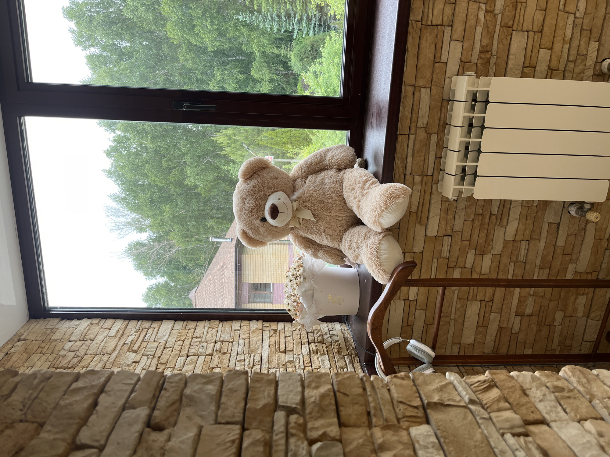 The image captures an interior scene looking out a window into a lush green landscape. The indoor space features a wall covered in light brown, textured rectangular stone tiles. A large window with a dark brown frame dominates the view.
On the wide windowsill or a shelf beneath the window, a large, fluffy light brown teddy bear with a white bow around its neck is seated. To its right, a white cylindrical box holds light-colored dried or artificial flowers. This box has some gold, stylized text and decorative motifs, which appear to read "Happy Day" or a similar positive phrase. Further to the right on the same surface, a pair of white headphones rests on a curved wooden stand.
To the right of the window, a white, finned radiator is mounted against the stone-tiled wall.
Outside the window, a dense canopy of green trees is visible, suggesting a forest or a heavily wooded area. Through the trees, parts of a yellow brick house with a brown roof can be seen in the distance.
The scene is set during the daytime, likely a bright but possibly overcast day, indicated by the diffused natural light. The overall atmosphere is cozy and domestic, set in what appears to be a residence with a natural view. There is no human activity depicted. The location context is Ореховка, Russia.