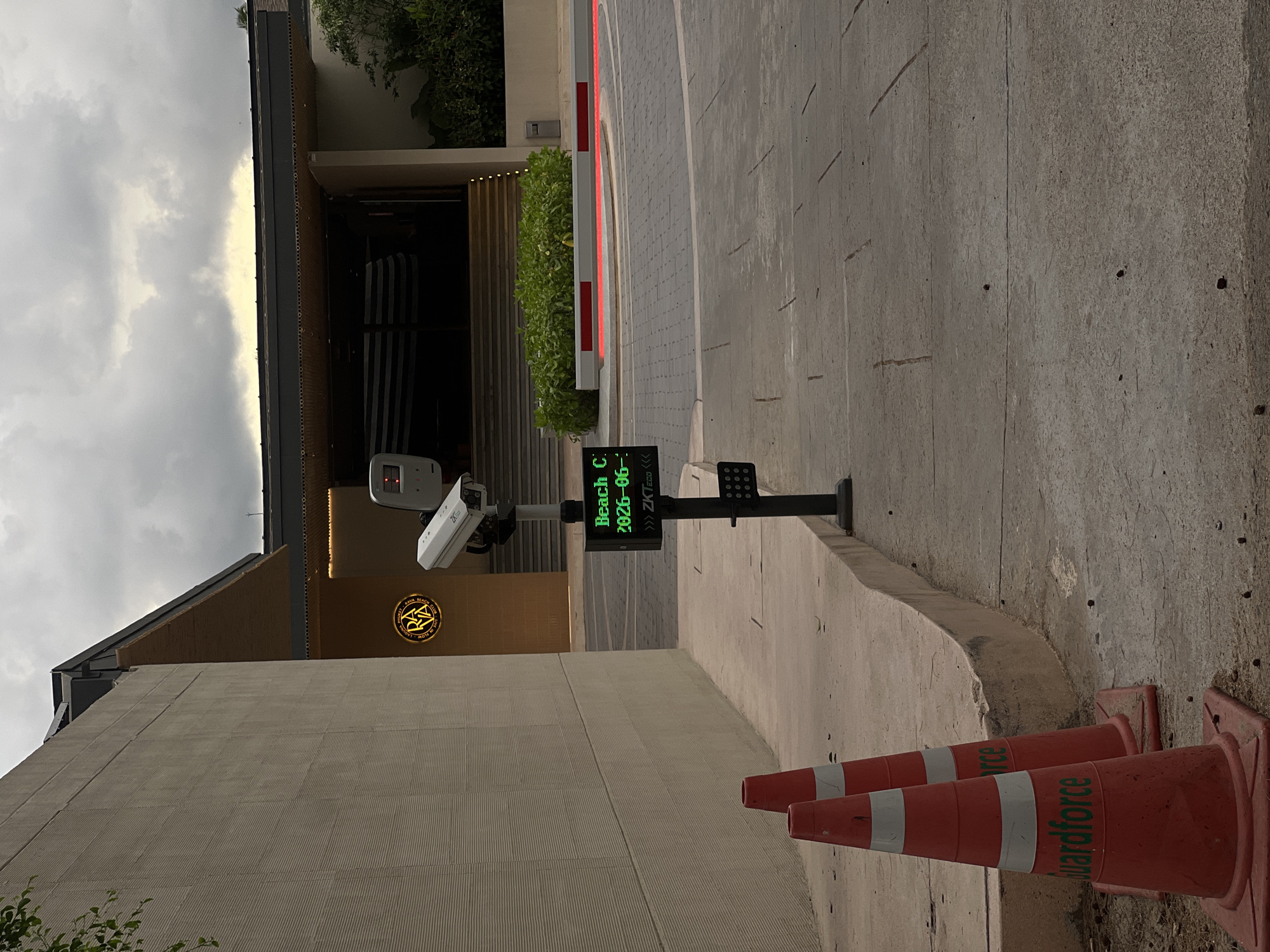 An outdoor scene in an unknown city shows a road leading to a building entrance. In the foreground, two orange traffic cones with white stripes are placed on a concrete surface. The left cone has "Guardforce" written in green text. To the right of the cones, a black pole supports a security camera and a digital display. The digital display shows "Beach C" in green text and "2026-06-" followed by a partially visible number. Below the display, a logo with "<< ZKTeco" is visible.
The building in the background has a light-colored textured wall on the left and a dark roof structure with recessed lighting over an entrance. A circular neon sign with the letters "RA" in yellow is mounted on the wall to the left of the entrance. A red and white barrier arm with red lights is extended across the road in front of the building. Green foliage is visible to the right of the entrance. The sky is overcast with clouds. The ground is paved with patterned concrete.