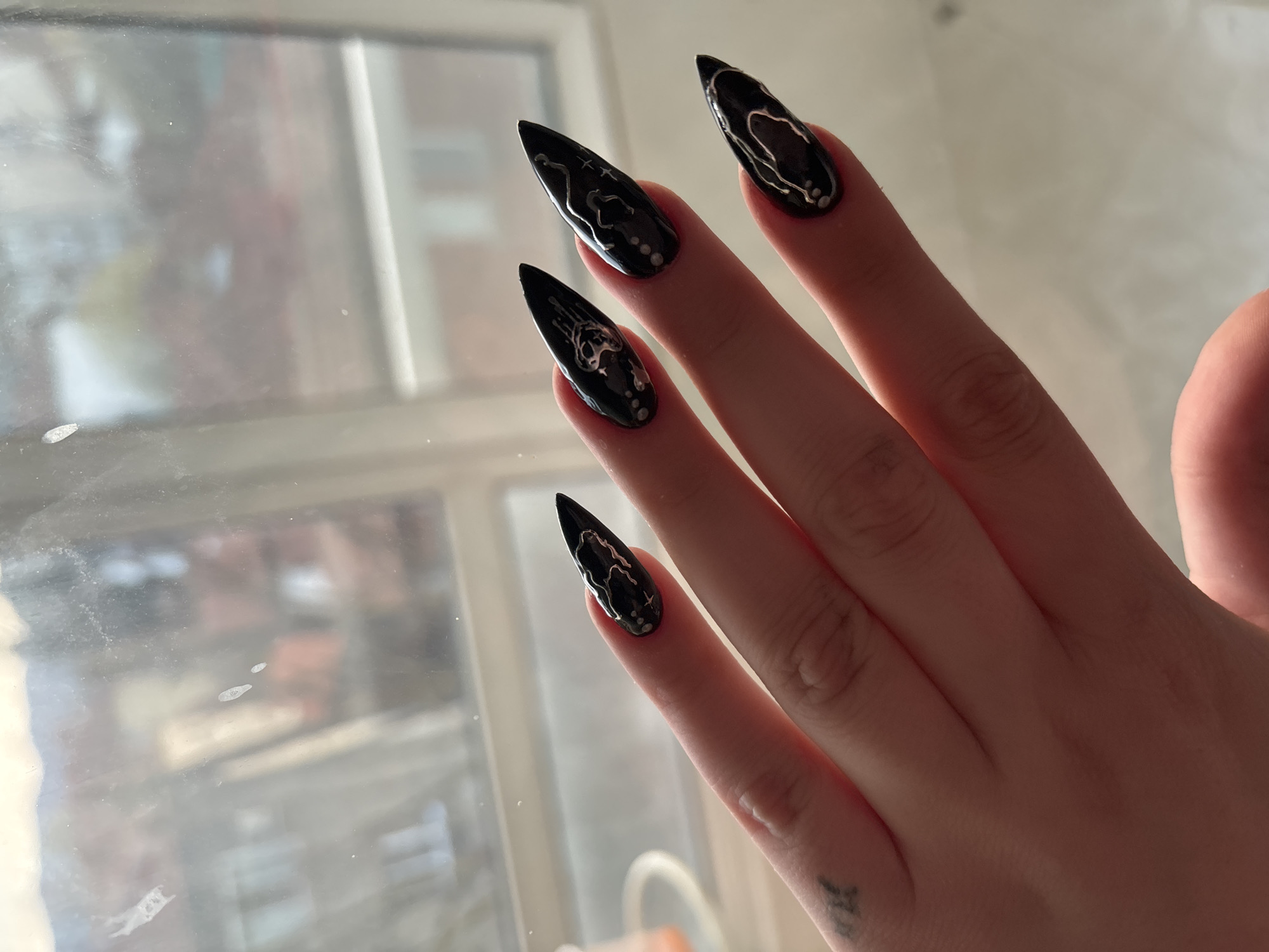 A close-up shot focuses on a person's hand, showcasing four elongated, stiletto-shaped nails painted with a glossy black base and intricate silver designs resembling veining or abstract patterns. Each nail is also adorned with small, silver-colored dots near the base. The hand is positioned against a blurred background, likely a window, partially revealing the urban environment outside with the window’s frame, including the outside scenery. A small, dark-colored tattoo is visible on the wrist. The location in the city of Troyan, Bulgaria is not evident.