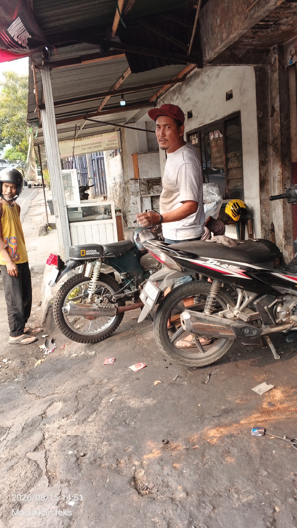 A street-side motorcycle repair shop is depicted in Malang, Indonesia, on August 15, 2026, at 14:51. Two men are present. One man, wearing a red cap and a white t-shirt, stands next to a motorcycle, appearing to work on it. He has a mustache and looks towards the camera. The second man, younger, wears a yellow t-shirt with graphics and a black helmet, standing to the left of the frame, looking away from the camera. Two motorcycles are the primary focus, with one being a dark green model with a National Geographic sticker on the fender and the other a newer model with red and white accents. The ground is dirty and appears to be a mix of dirt and asphalt, with scattered debris, including what looks like small pieces of paper. The background shows the facade of a building, possibly a workshop, with open doors and shelves. A yellow helmet rests on a surface behind the motorcycles. The environment suggests a typical urban setting in Indonesia, with the presence of motorcycles indicating common transportation.