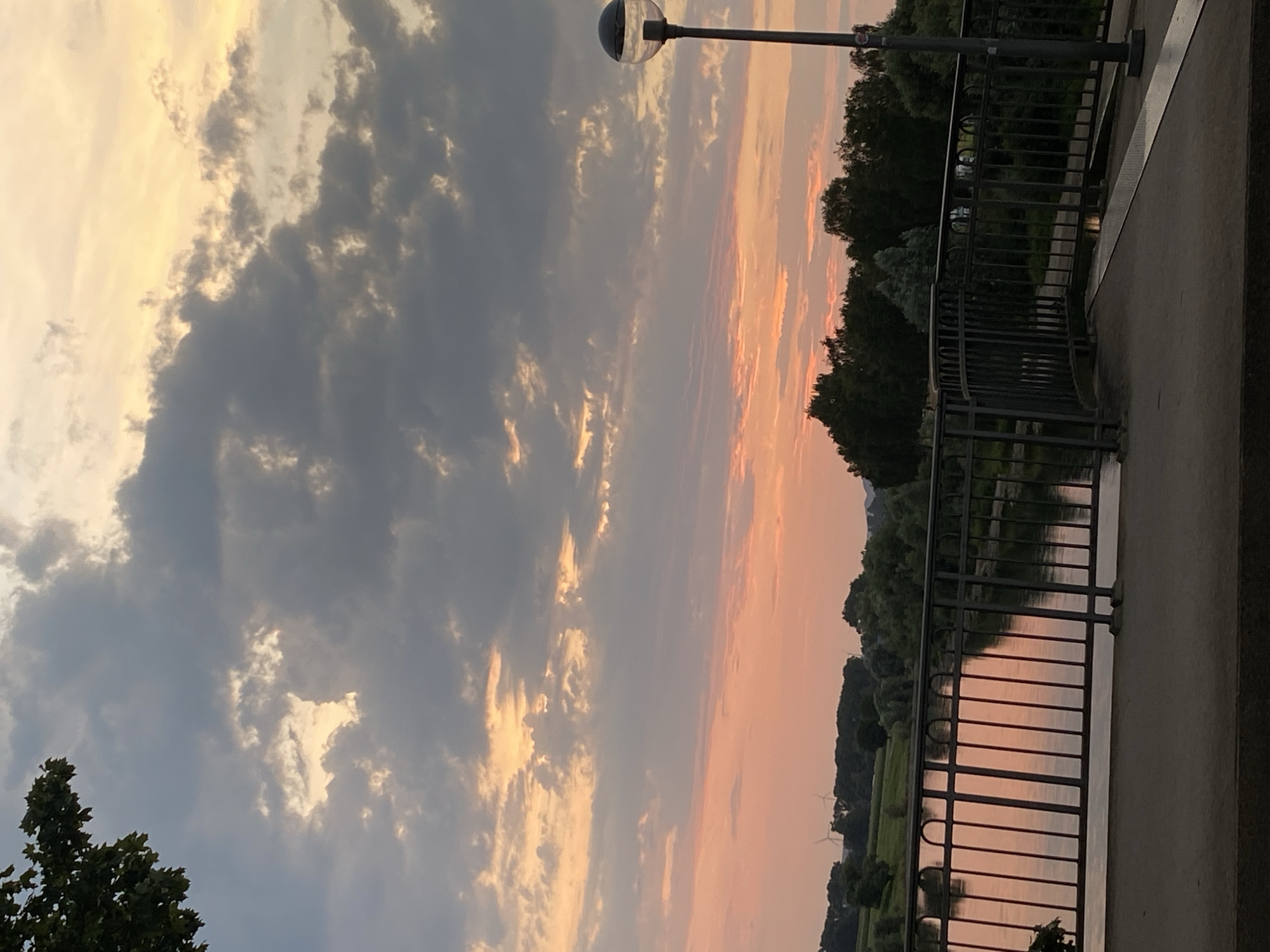 An elevated view captures a sunset over a river in Rinteln, Germany, with a modern lamppost in the foreground on the right. Dark, textured clouds are prominent in the sky, with streaks of orange and yellow light indicating the setting sun. Below the clouds, the sky transitions to a soft pink hue. A metal railing of a bridge or elevated walkway runs across the middle of the frame, with a river reflecting the sunset's colors. The riverbank is lined with dark green trees, and in the distance, a few wind turbines are visible on a grassy field. The pavement of the walkway is visible in the bottom of the frame.