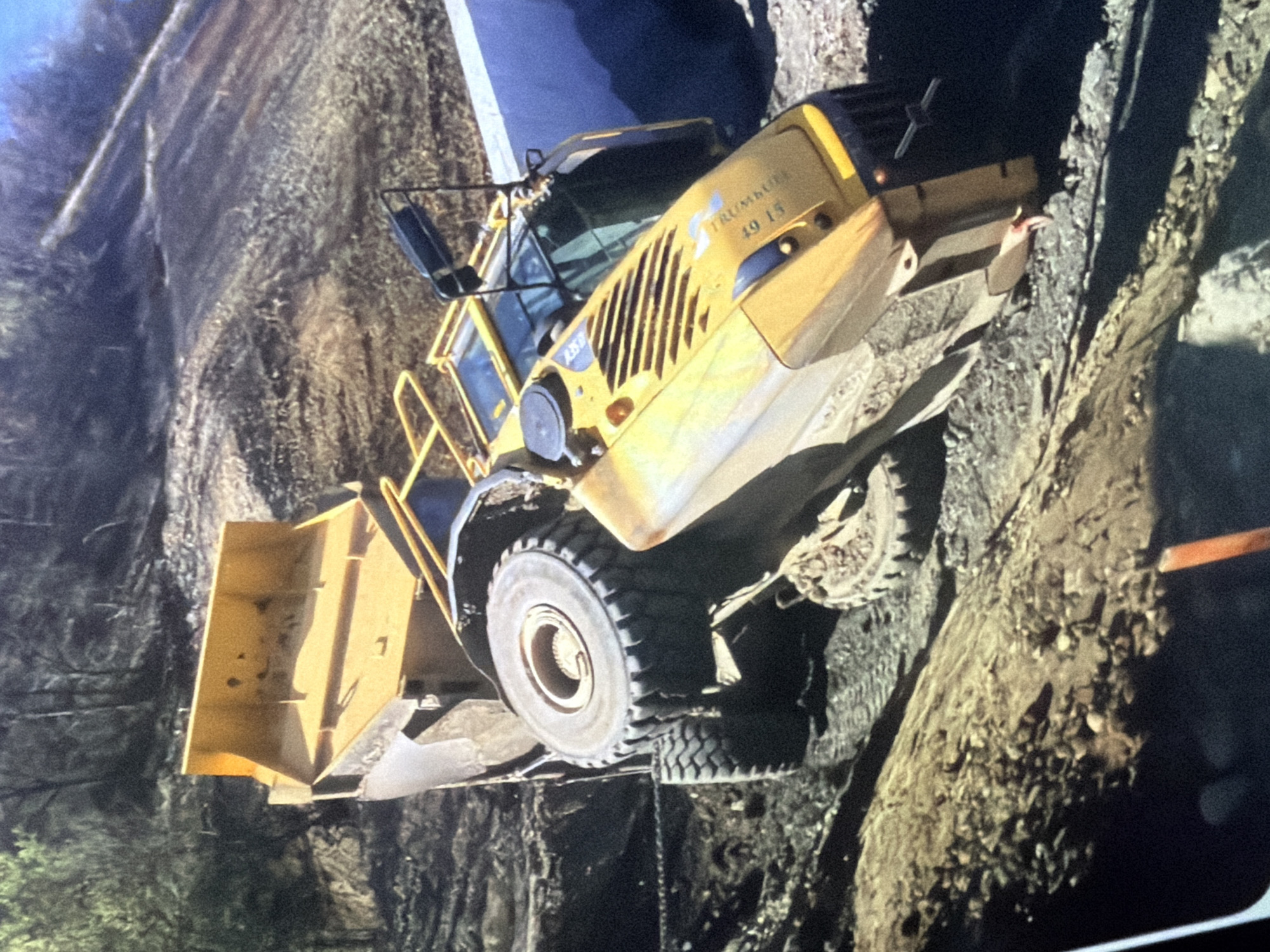 An overturned yellow construction vehicle, possibly a dump truck, is depicted in a dirt quarry. The vehicle is on its side, with one of its large rear tires still visible. The front of the vehicle, including the cab and engine compartment, is angled upwards. Debris and loose dirt surround the vehicle, suggesting a recent accident or collapse. The background shows a wooded area on a slope. The location is identified as a quarry in Unknown, Unknown.