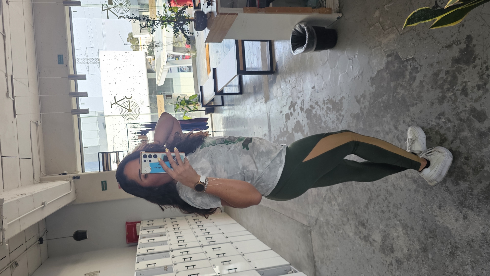 A woman is taking a mirror selfie inside a building. She is wearing a white t-shirt, green and tan leggings, and white sneakers. She has dark hair and is holding a smartphone in front of her face. The building appears to be a gym or fitness center. The location is in the city of Puebla, Mexico. The lockers, plants, and furniture provide evidence of this.