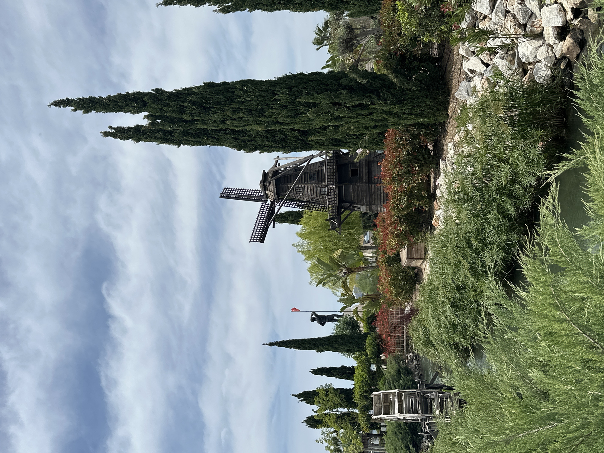 The image depicts a picturesque scene in Fethiye, Turkey, featuring a wooden windmill, a statue, and surrounding foliage.
The windmill is centrally positioned and appears to be an older, weathered structure. Its dark wood construction contrasts with the greenery around it.
In the distance, a statue can be seen, possibly depicting a figure in mid-action, standing atop a platform with a red flag waving in the background.
The environment includes a variety of trees and bushes, framing the view with green and brown tones. A small water feature is at the bottom of the image, reflecting some of the elements above. The sky is partially visible, showing a mix of clouds and clear sky. The overall scene has a park-like or garden-like setting.