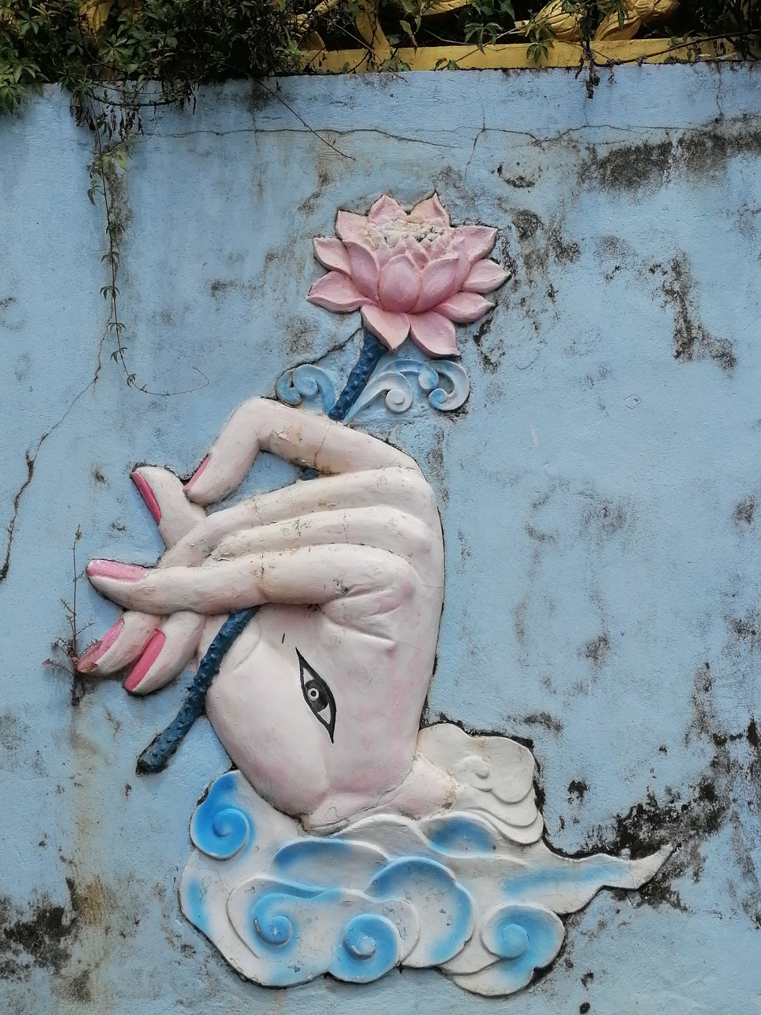 The image shows a bas-relief sculpture on a light blue, weathered wall in Da Lat, Vietnam. The sculpture depicts a stylized hand with pink fingernails holding a pink lotus flower. The hand is detailed with visible lines and texture, and one eye is painted onto the palm area. The hand emerges from a decorative element resembling stylized white and blue clouds. Green vines hang from the top of the wall, partially obscuring the upper edge and casting shadows. The wall itself shows signs of age, with cracks and dark staining. The overall aesthetic suggests a decorative element, possibly from a temple or garden setting in Da Lat.