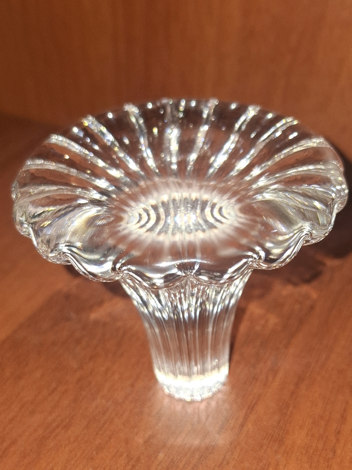 The image shows a clear glass object with a fluted, petal-like design on top and a fluted stem. The object is placed on a wooden surface. The background is a warm, brown tone. The lighting creates reflections and highlights on the glass. This image is not located in the city of Almaty, Kazakhstan.