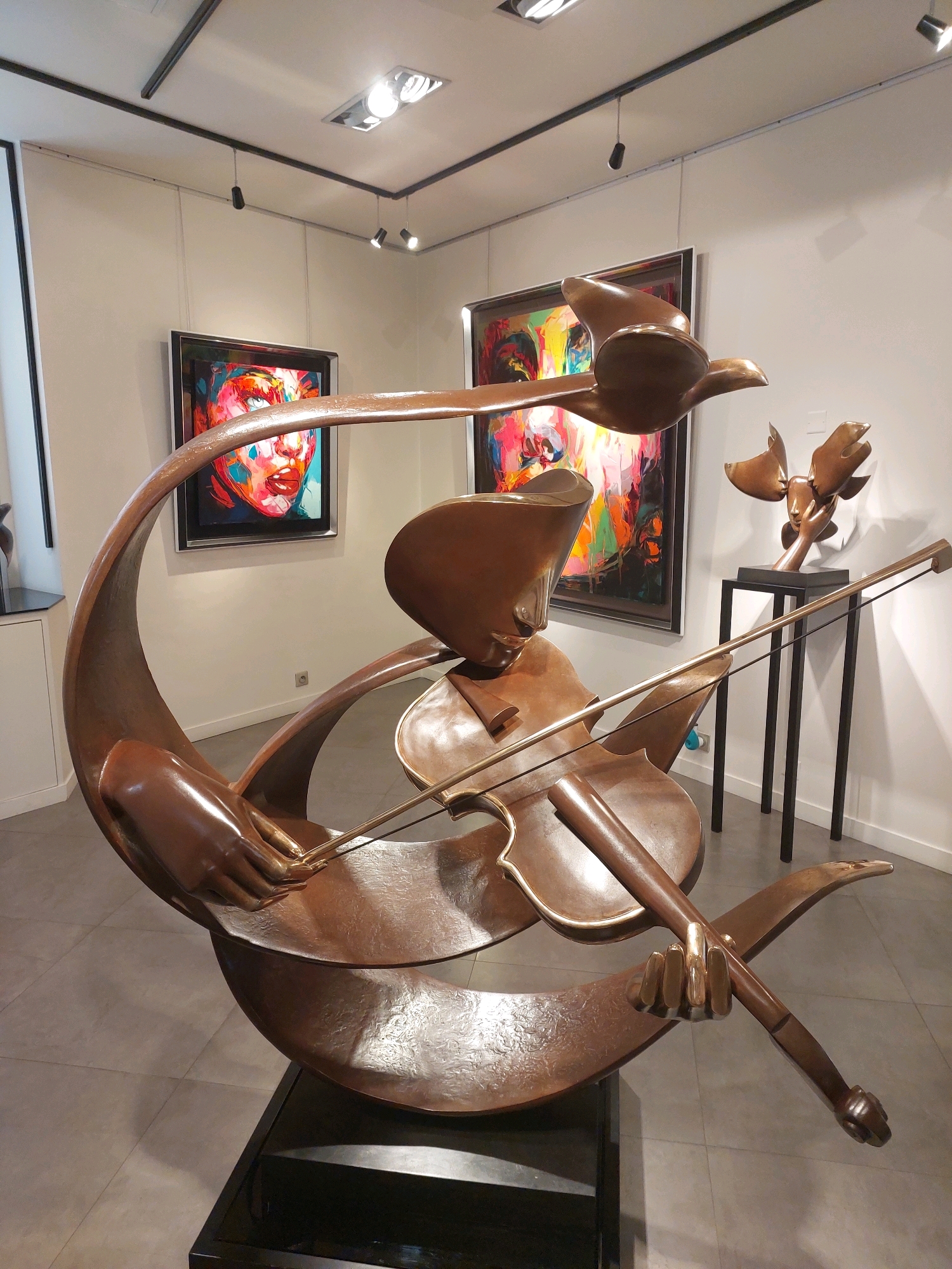 The image depicts a bronze sculpture of a figure playing a violin, with stylized birds in flight. The sculpture is located in a gallery in Saint-Paul-de-Vence, France. The sculpture is abstract, with smooth, flowing lines and a warm bronze finish. The figure is depicted with a serene expression, and the birds appear to be soaring freely. The background of the image shows a brightly lit gallery space with other artworks on display, including a colorful abstract painting. The floor is tiled, and the walls are painted white. The overall impression is one of artistic sophistication and a celebration of nature and music.