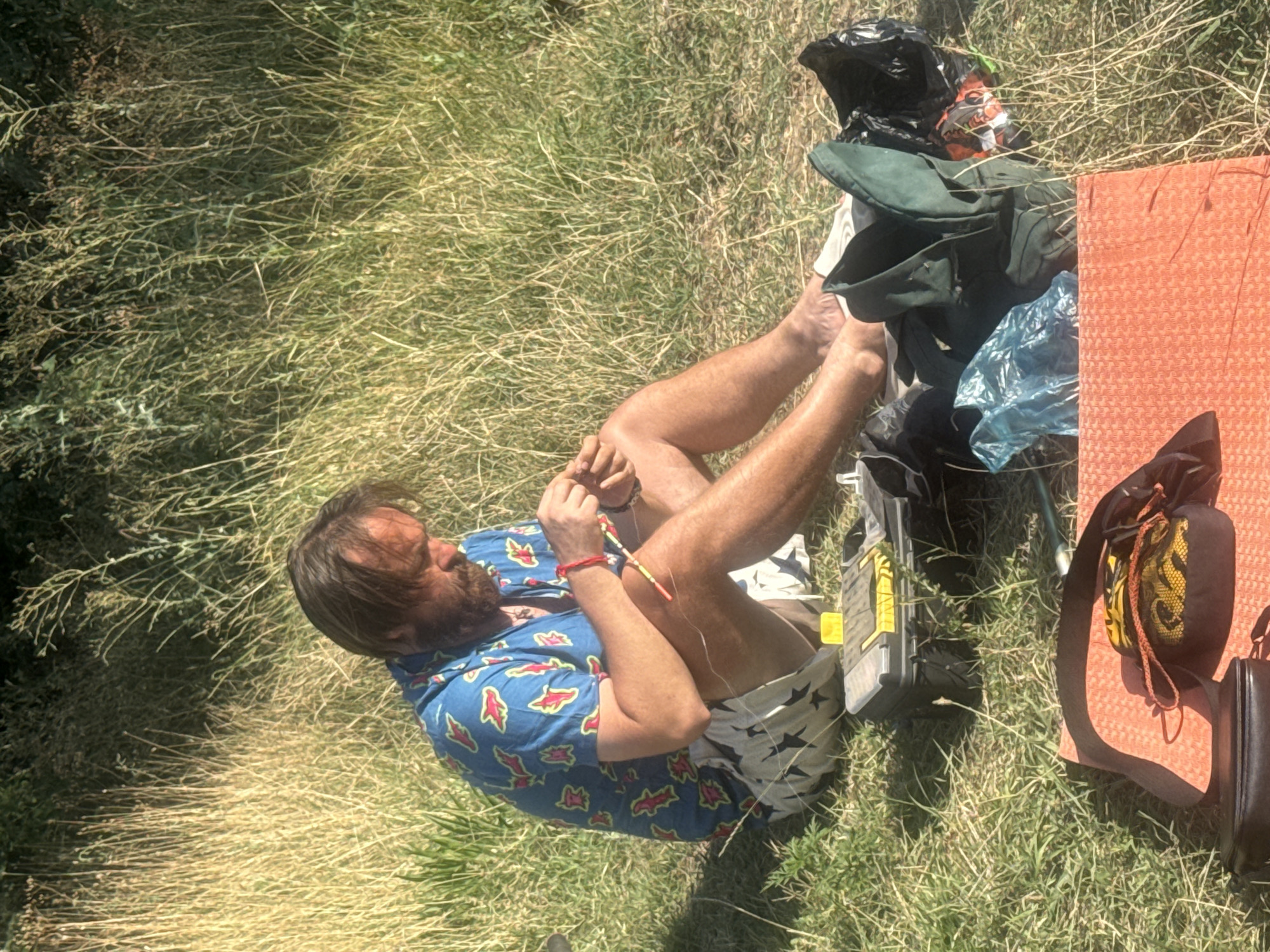 The image depicts a man sitting on the ground in a grassy, outdoor environment, likely in Муромское сельское поселение, Ukraine. He is wearing a blue shirt with a colorful pattern and patterned shorts. The man is focused on an activity involving small objects, possibly crafting or repairing something with his hands, as he holds a string of beads. Around him, there are various items including a fishing tackle box, a dark bag, a green bag, a blue plastic bag, and an orange mat. The background consists of tall dry grass and some foliage, suggesting a rural or park-like setting. The lighting indicates it is daytime, with sunlight casting shadows.