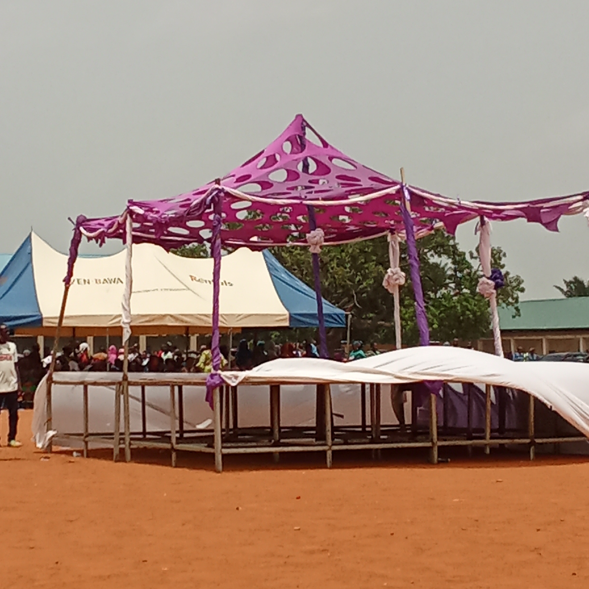 The image depicts an outdoor event or market scene, likely set up on a reddish-brown dirt ground. In the foreground, a vibrant temporary canopy or stall features a unique design with purple fabric adorned with circular cutouts, supported by poles. White fabric is draped around and beneath this structure, covering what appear to be raised tables or stands, possibly for displays or serving.
In the mid-ground, behind the foreground setup, a gathering of people can be seen, partially obscured. They appear to be seated or standing, forming a crowd. Two additional tents are visible further back. One is a large white marquee tent with blue trim, on which the partial text "EN BAWA" and "Rentals" is visible, suggesting it's rental equipment for events. Another blue tent is situated to its right.
The background shows scattered green trees and a building with a green roof. The sky is bright and appears overcast or hazy, indicating daytime. A person wearing a white t-shirt and dark trousers is seen walking on the far left of the frame. The overall scene suggests a communal gathering or public event taking place in an open area. The setting aligns with the provided context of Akwanga, Nigeria, with the reddish soil being a notable environmental cue.