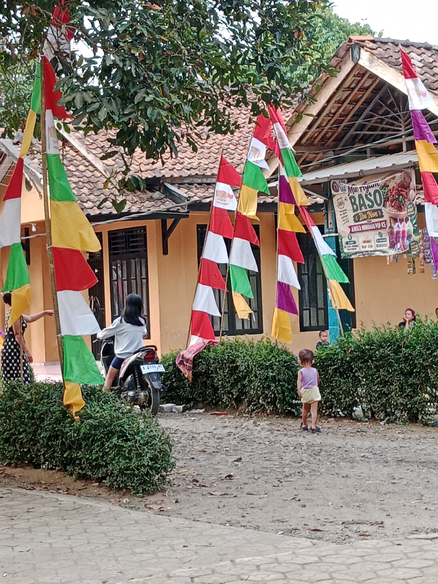 In Parage, Indonesia, a street scene unfolds with a row of houses and lush green hedges. Indonesian flags in red, white, green, and yellow flutter on poles, some adorning the houses. A woman in a polka dot dress stands to the left, pointing towards a young girl on a motorcycle. The girl, wearing a white sweater and dark shorts, sits on the motorcycle, facing away from the camera. A toddler in a purple top and yellow shorts stands near the hedges on the right, looking away from the viewer. A banner advertising "Mie Nyum BASO" and "IBU NON" is visible on one of the houses. The ground is a mix of dirt and paving stones.