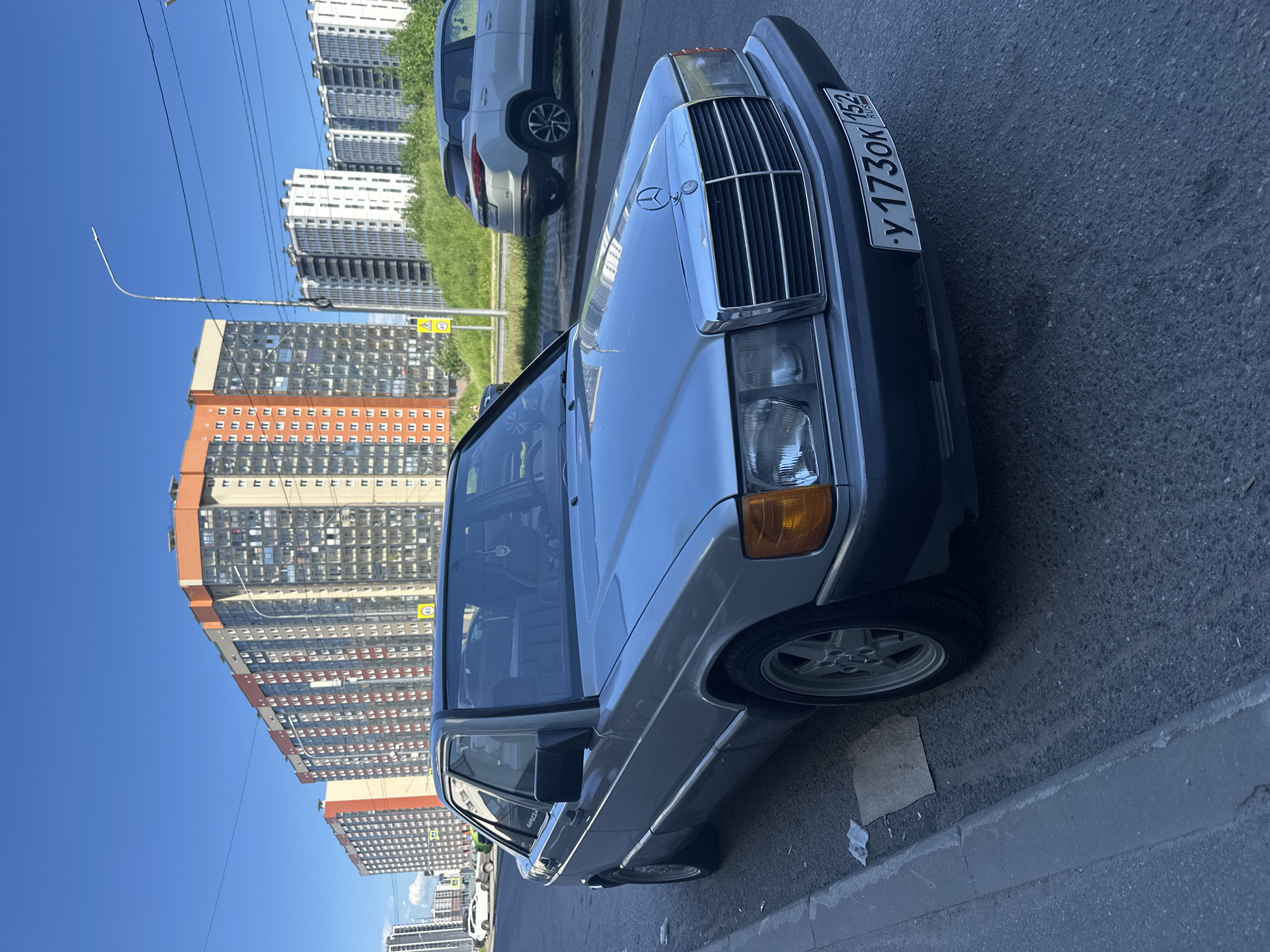 A silver Mercedes-Benz 190E is parked on the side of a street in Murino, Russia. The car is positioned facing the right of the frame, with its front end visible. In the background, several modern high-rise apartment buildings stand under a clear blue sky. A white SUV is parked on the opposite side of the street, further down. The street is paved with asphalt, and there is a concrete curb alongside the parked Mercedes. A small piece of cardboard and some white litter are on the ground near the curb. The license plate of the Mercedes reads "y 173ok 152 RUS".