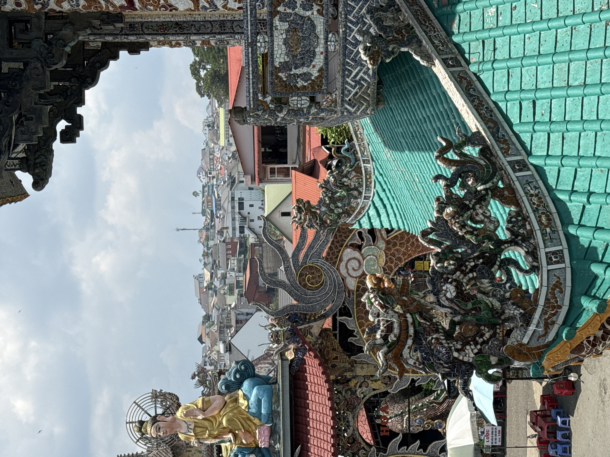 This image shows a view in Xuân Thành, Vietnam, likely taken from a temple or ornate structure. The foreground is filled with elaborate architectural details, featuring mosaics, sculptures, and green tiled roofing. A golden statue is visible at the bottom left. Beyond the structures, a dense cityscape stretches into the distance under a partly cloudy sky. The buildings are primarily residential, with a mix of roof colors.