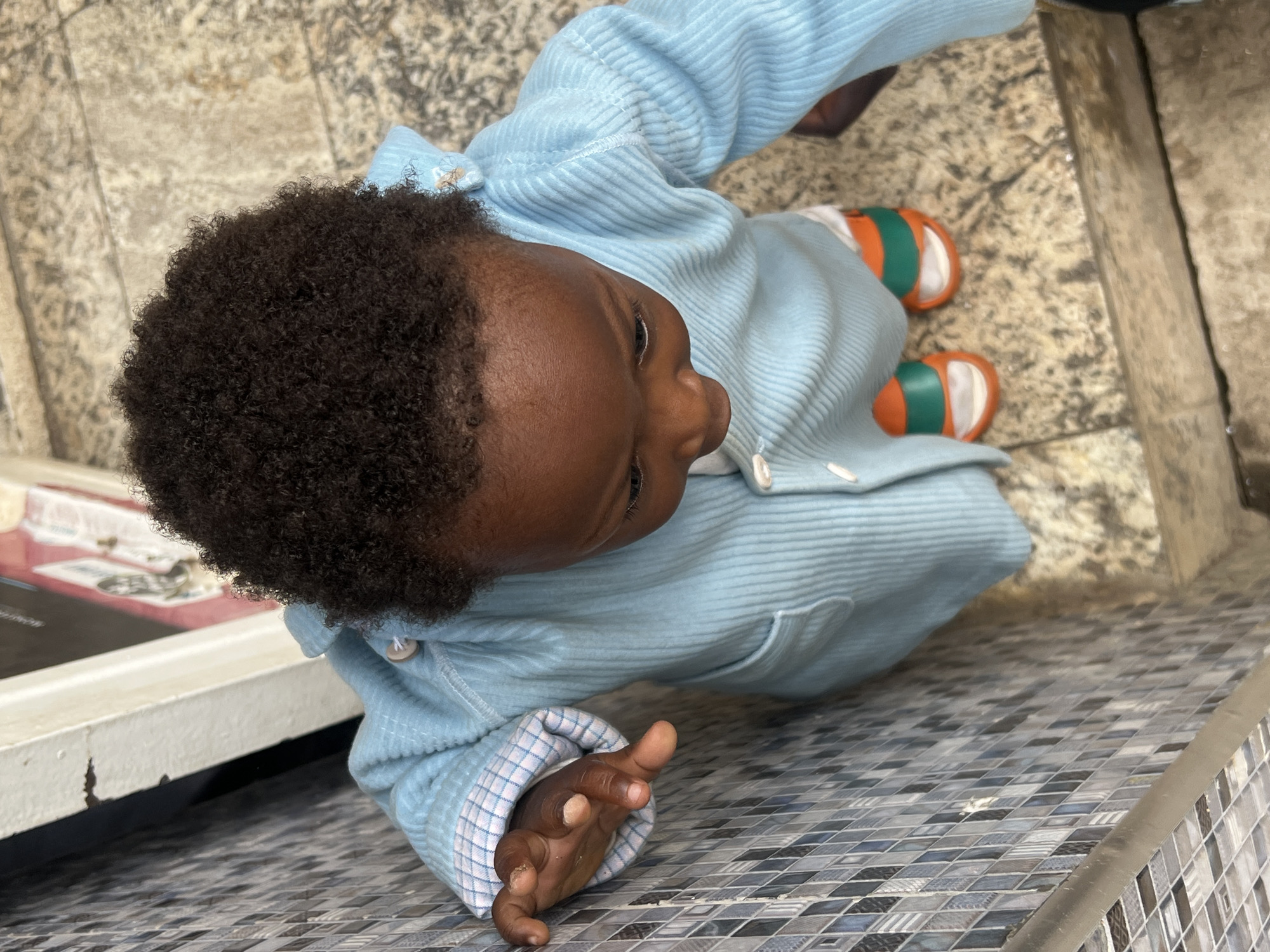 A small child with dark skin and tightly curled black hair is seated on a tiled surface. The child is wearing a light blue button-down shirt with checkered cuffs and orange and green sandals. The tiled surface appears to be a step or ledge. The location is likely Gura Top, Nigeria, as specified in the prompt, but there are no specific indicators in the image to confirm the exact location within the city.