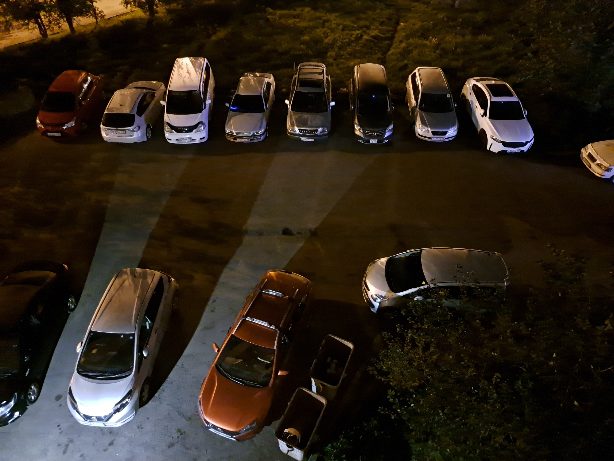 An overhead drone shot captures a dimly lit, outdoor parking lot at night in Irkutsk, Russia. Several cars are parked in neat rows. On the upper left, a red hatchback is parked next to a white compact car. To the right of the white car is a silver sedan, followed by a darker sedan, and then two lighter-colored sedans. Further to the right, a white station wagon is parked. In the lower portion of the frame, a black SUV is parked next to a silver hatchback, followed by an orange sedan. Two light-colored rectangular objects, possibly trash bins, are situated near the orange sedan. The parking lot is paved and appears wet, reflecting some of the ambient light. Adjacent to the parking lot, to the left and top, are trees with sparse foliage illuminated by an unseen light source. The overall impression is a quiet, nocturnal scene in a residential or commercial area of Irkutsk.