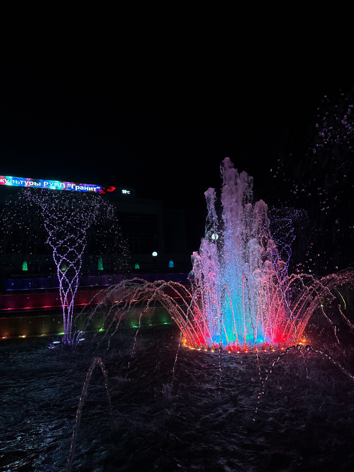 This is a night-time, low-angle shot of a vibrant, illuminated fountain in the city of Mikashevichy, Belarus. The fountain features multiple streams of water, with the central and largest jet illuminated in bright blue, pink, and red hues. Surrounding this central jet are smaller arcs of water, also colored with pink and red lights. The water splashes into a dark pool, creating ripples and reflections of the lights. In the background, a building facade is visible, topped with a brightly lit sign in Russian that reads "культуры РУПП 'Гранит'" (Culture RUPP "Granit"). To the right of this sign, a digital display shows "14°C". Several smaller, cone-shaped lights in green, blue, and yellow are embedded in the tiered structure of the fountain base, adding further color to the scene. The overall environment is dark, with the fountain and sign being the primary light sources. No people are visible in the frame.