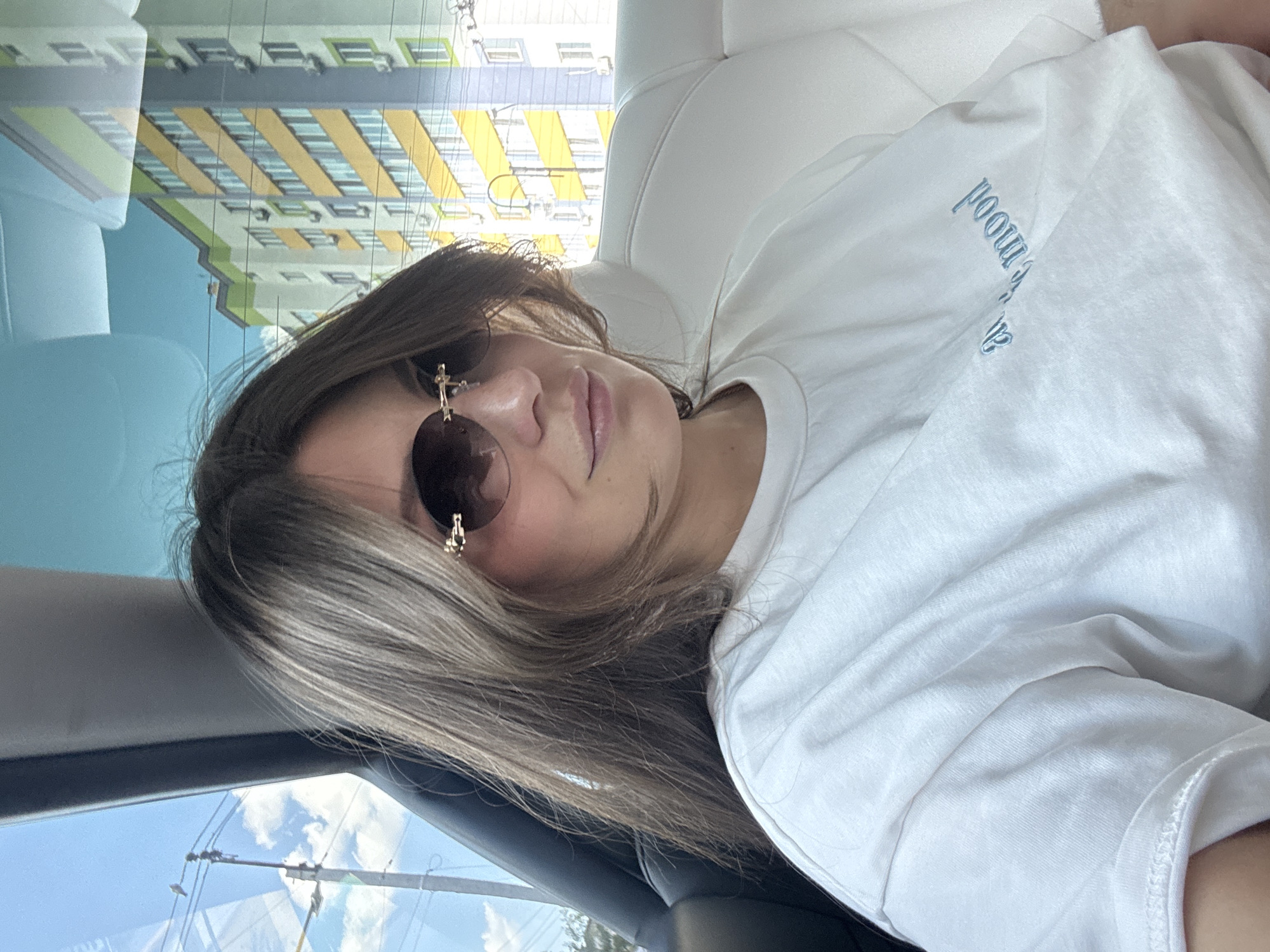 A young woman is captured in a relaxed pose, likely reclining in the passenger seat of a vehicle. Her head is tilted, and she looks directly at the camera, wearing large, dark sunglasses adorned with distinctive gold-colored cross-like embellishments on the temples. Her medium-length, light brown hair with blonde highlights frames her face. She is wearing a simple white t-shirt, on which light blue embroidered text is visible. When viewed correctly, the text reads "in the mood."
The setting appears to be daytime with bright, sunny weather, indicated by the clear blue sky and white clouds visible through the car window. Reflections and views through the window reveal an urban or suburban environment. A prominent multi-story residential building with yellow and light green striped facade is reflected in the upper part of the window, suggesting a modern architectural style. Utility lines are also visible against the sky. The location context is Sofiivska Borshchahivka, Ukraine. The overall mood of the image is calm and casual.
