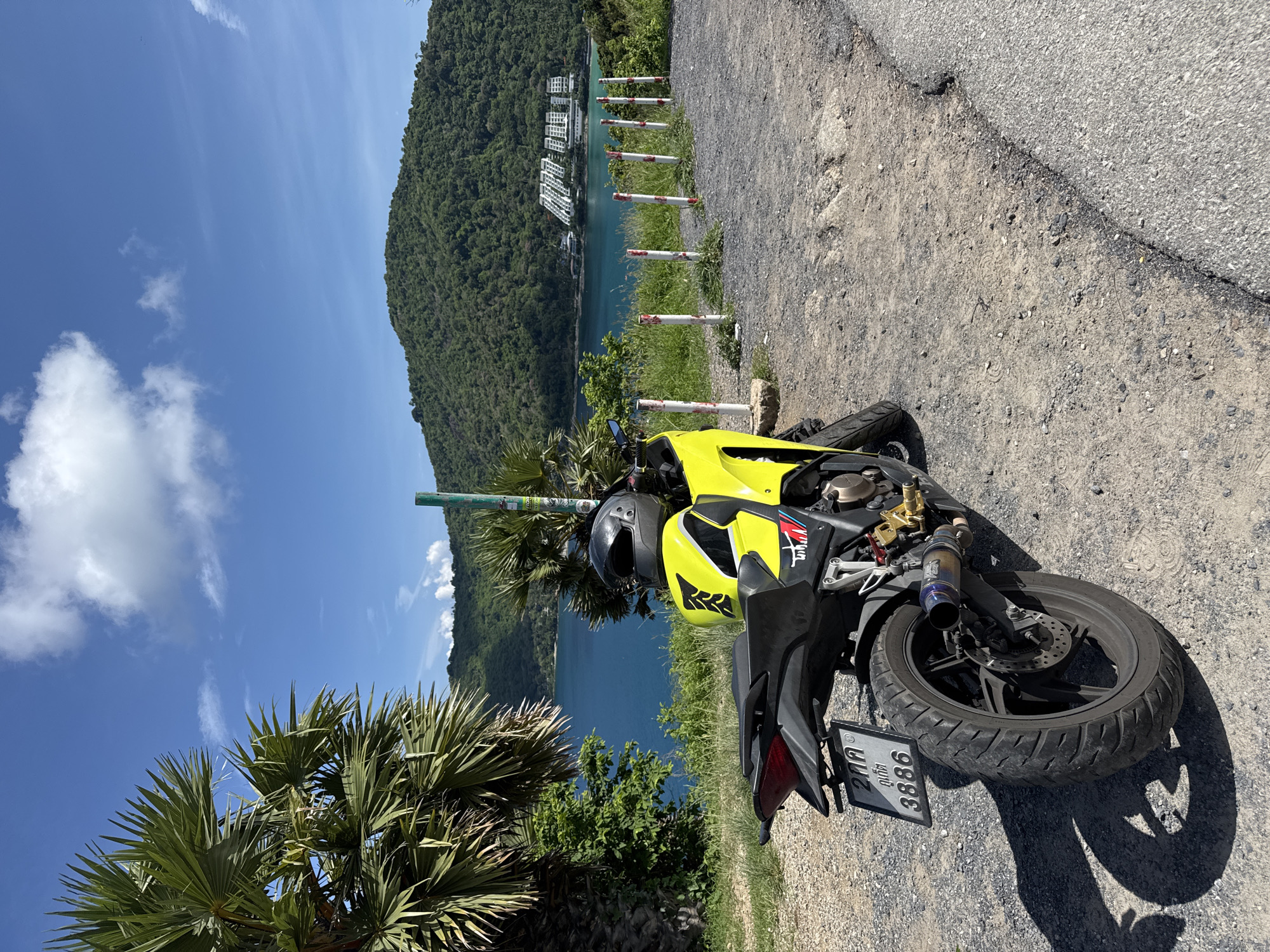 In an outdoor, daytime scene, a yellow and black motorcycle is parked on a gravelly surface next to a paved road. The motorcycle is facing away from the viewer, with its rear wheel and tail prominently displayed. A black helmet rests on the seat of the motorcycle. In the background, a body of turquoise water is visible, bordered by lush green hills covered in trees. Several white posts with red tops are lined up along the edge of the gravel area. A large palm tree with fronds is on the left side of the frame. The sky above is blue with scattered white clouds. The location is described as Unknown, Unknown.