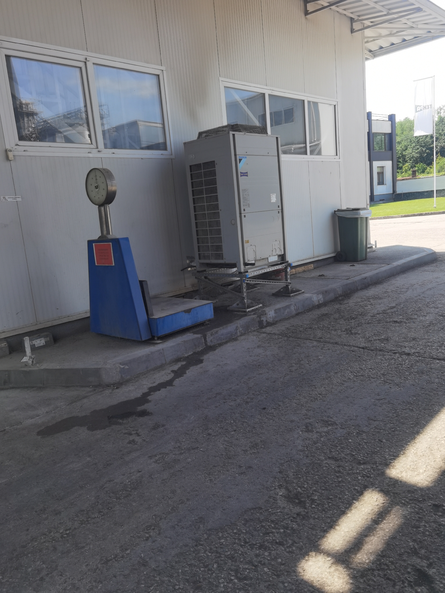 An outdoor scene is captured in Lomets, Bulgaria, showing a large industrial building with a white, ribbed facade and several windows. In front of the building, there is a blue weighing scale with a circular dial. Next to the scale, an industrial air conditioning unit is mounted on a metal stand. A green trash bin is situated behind the air conditioning unit. The ground is paved with asphalt and concrete, with a slight slope. In the background, trees and a white flag with text are visible, suggesting a commercial or industrial area. The lighting indicates a bright, sunny day.