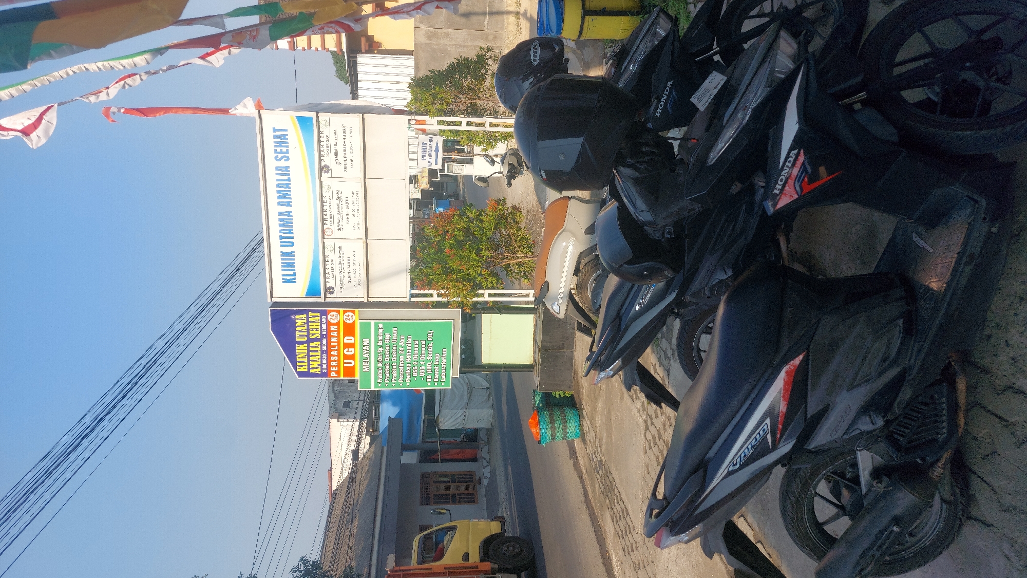 The image shows a street scene in Sedan, Indonesia, with the main focus on a clinic. A sign for "KLINIK UTAMA AMALIA SEHAT" is prominently displayed, with information about services like obstetrics and gynecology (PERSALINAN) and a 24-hour emergency unit (UGD). Another sign to the right lists doctor's practices and their schedules. In the foreground, several motorcycles are parked, some with helmets on them. In the background, a building that appears to be part of the clinic is visible, along with some trees and a street with a yellow truck. The sky is clear and blue. The overall impression is of a functional medical facility on a sunny day in a semi-urban or town setting.