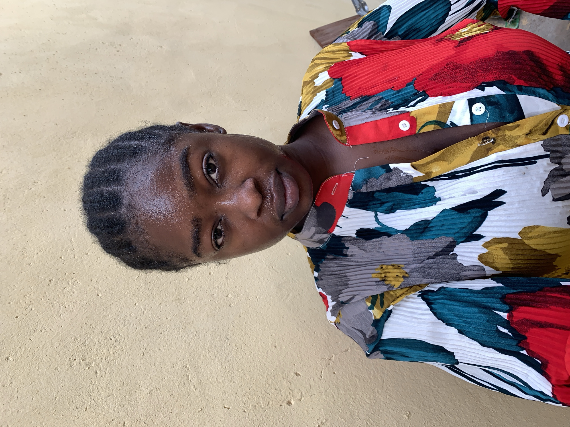 The image shows a dark-skinned woman with braided hair wearing a colorful, floral-patterned shirt. She is positioned against a plain, light yellow wall. The location, Rafin Par, Nigeria, is not verifiable from the image.