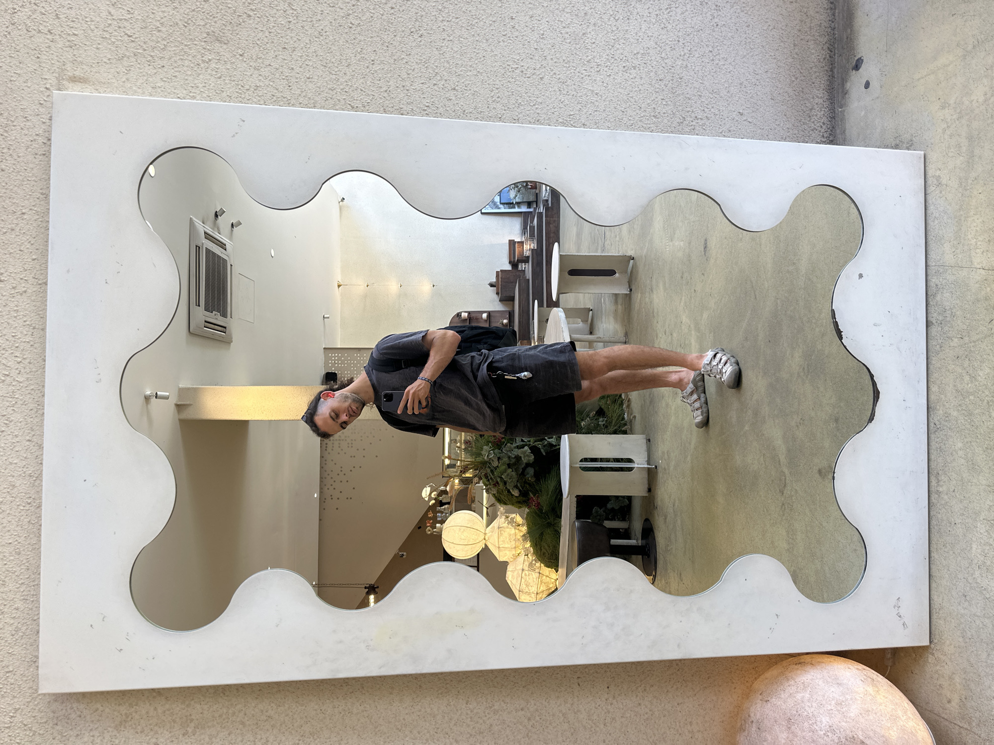 A young man with dark, curly hair tied back in a bun is taking a selfie in a large, irregular-shaped mirror. He is wearing a dark grey t-shirt and dark shorts, with grey sandals. He is holding his phone in his right hand, and a backpack is visible on his back. In the reflection, the man is standing in what appears to be a cafe or retail space with a concrete floor and beige walls. There are various interior design elements visible, including modern furniture, decorative lighting fixtures, and plants. The overall ambiance is bright and contemporary. The scene is located in Đà Nẵng, Vietnam.