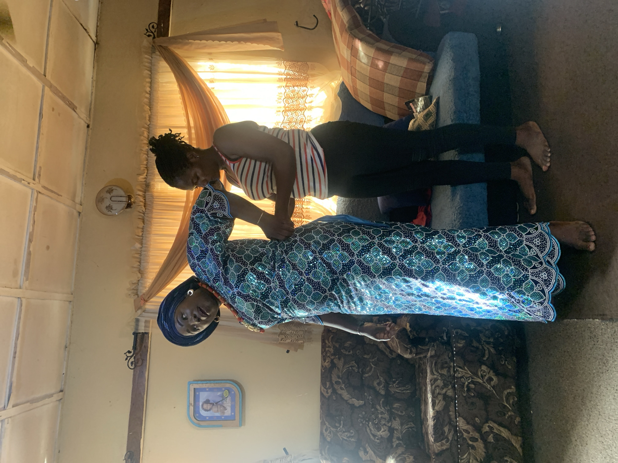Two women are in what appears to be a living room. One woman, dressed in a long blue patterned dress and a headwrap, is kneeling on the floor while the other woman, in a striped tank top and black leggings, is lying on her back on a blue sofa. The kneeling woman is supporting the back of the other woman. The room has beige walls, and a sheer curtain is visible in the background. A small framed photo is on the wall. There's a patterned cushion on the sofa. The location is Rafin Bauna, Nigeria.
