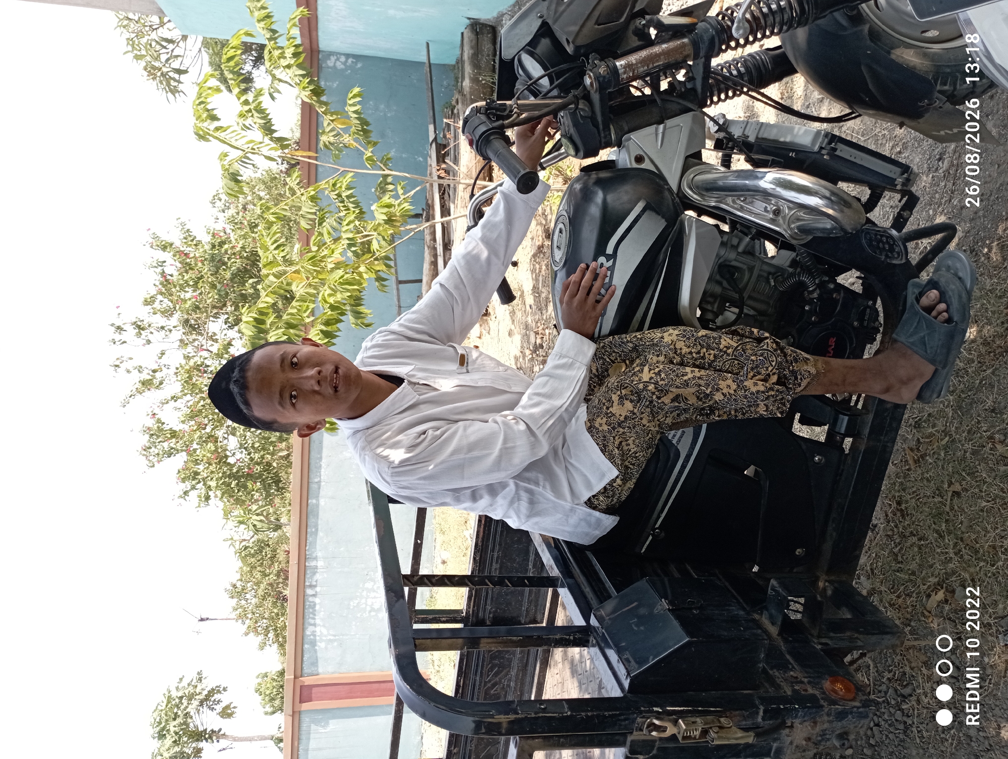 A young boy wearing a white shirt, patterned pants, and a black skullcap sits on a black and silver motorcycle with a sidecar attachment. The motorcycle is parked on a dirt and grass surface. The boy is looking directly at the camera. In the background, there is a light blue and pink wall, trees with green and some red leaves, and a bright sky. The image also has timestamps indicating it was taken on August 26, 2026, at 13:18, and a Redmi 10 2022 watermark. The scene appears to be outdoors in a rural or suburban area, possibly in Kecamatan Pati, Indonesia, given the timestamp and context.