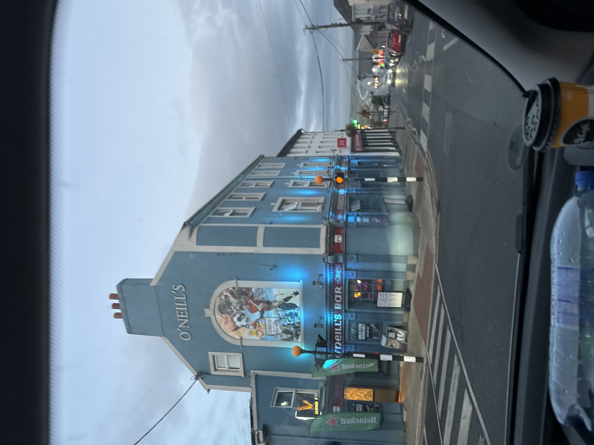 The image shows a street scene in Tramore, Ireland, viewed from inside a vehicle. The primary focus is on a light blue building with "O'NEILL'S" signage and "O'NEILL'S BAR" below. The building is illuminated with blue lights, and there is a mural on the upper facade depicting a sports scene. Green banners with "Heineken" branding are visible on the left side of the building, along with a Guinness logo. A traffic light is visible in front of the building. The street is wet, suggesting recent rain, and there are parked cars and other vehicles further down the road, some of which have their headlights on. The sky is overcast. A plastic bottle and a McCafe cup are in the foreground, on the dashboard of the vehicle.