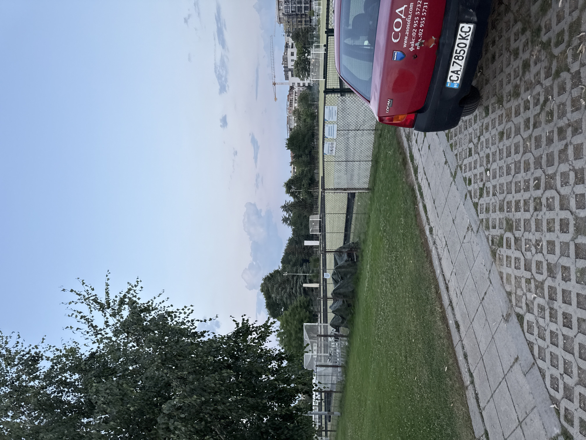 The image shows a red Dacia Logan car parked on a paved surface in front of a green lawn. Beyond the lawn is a fenced sports field, likely a football pitch, with goalposts visible. In the background, there are trees and buildings under construction, including a crane. The sky is a pale blue with some scattered clouds. The car has Bulgarian license plates with the registration number CA 7850 KC. There are logos and text on the car's trunk, including "DACIA", "LOGAN", and "COA" with a website address and phone numbers. The overall scene suggests an outdoor location in Sofia, Bulgaria, possibly during the late afternoon or early evening.