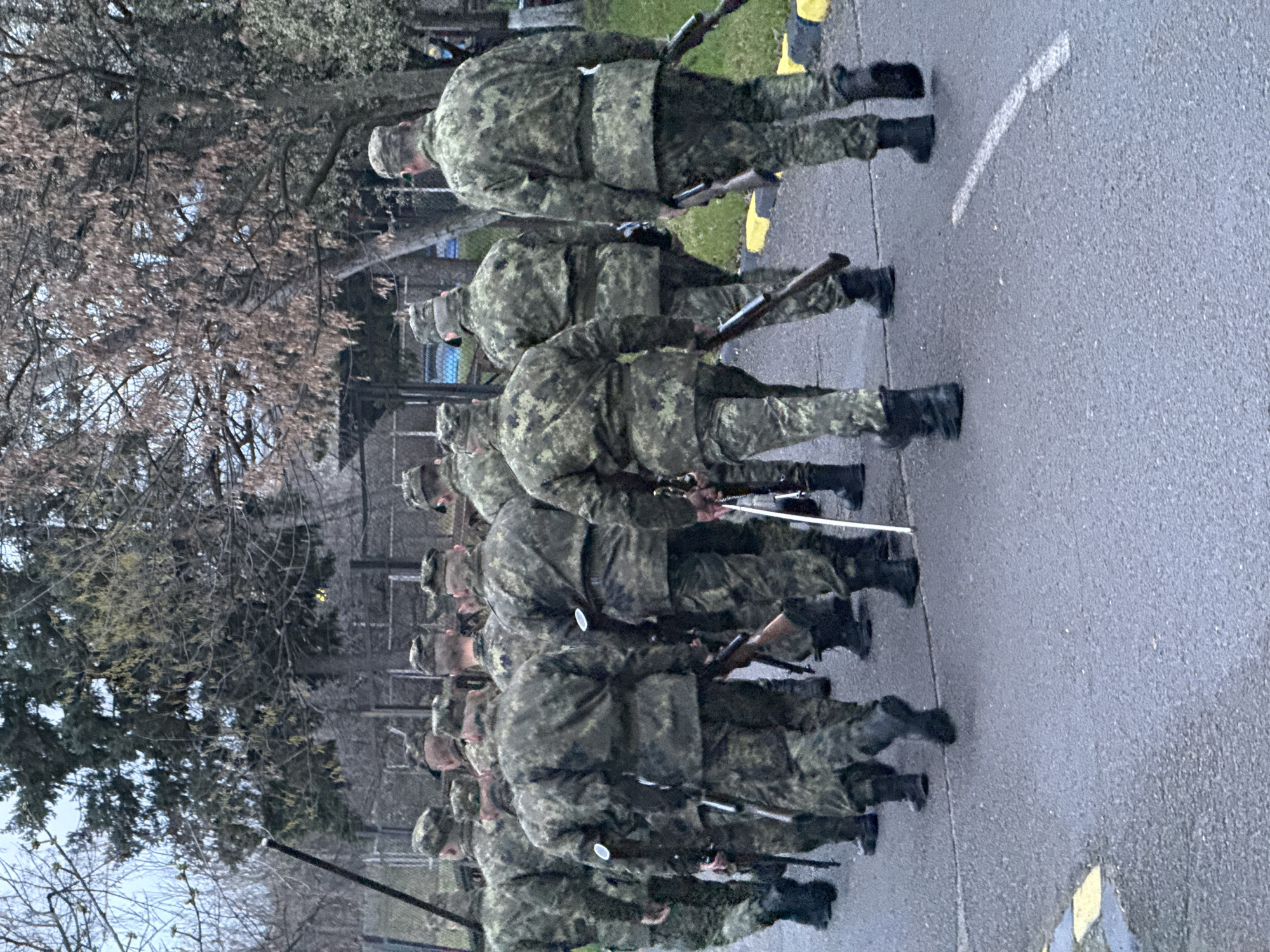 A line of soldiers in camouflage uniforms and black boots stands in formation on an asphalt surface, possibly a road or parade ground. They are holding what appear to be rifles and sabres. In the background, there are bare trees and the edge of a building. The location is Sofia, Bulgaria.