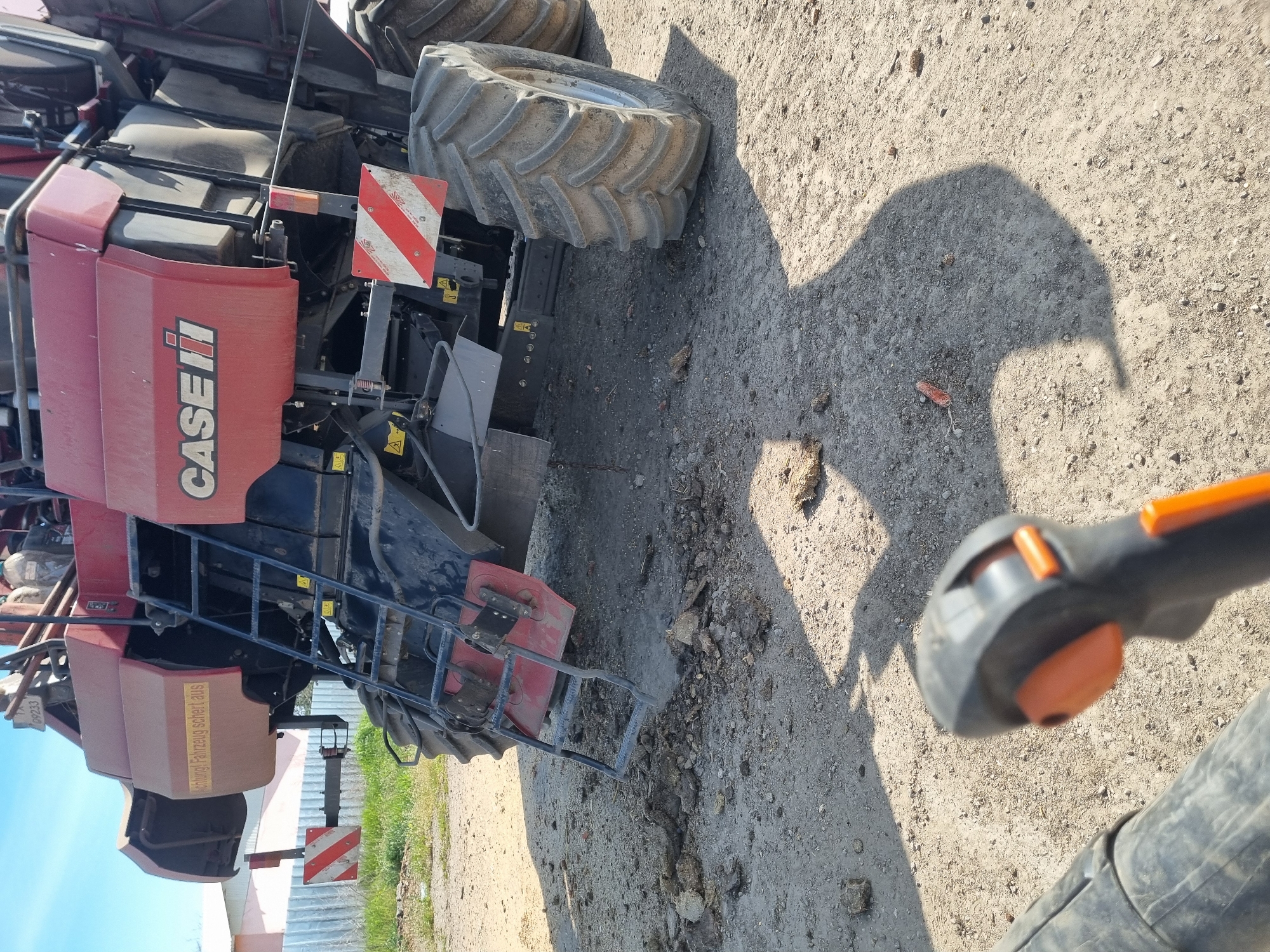 A large red and black combine harvester, branded "CASE IH," is parked on an unpaved, dusty, and rocky surface, likely a farmyard or rural road in Ovcharovo, Bulgaria. The machine is equipped with massive, heavily treaded tires and a black metal ladder for access. A red and white striped warning sign, typical for oversized agricultural vehicles, is affixed to its side.
In the lower right foreground, the orange and black handle of a leaf blower or similar cleaning device is partially visible, indicating a potential cleaning or maintenance activity. A distinct shadow, likely cast by a person or object, stretches across the dusty ground, suggesting bright sunlight. The ground is littered with small rocks and dried earth debris.
The scene is set outdoors under clear, sunny skies, with strong shadows indicating either morning or late afternoon. In the background, a light-colored fence or building structure can be partially seen.
Visible text includes the "CASE IH" logo on the side of the harvester. A yellow warning sticker on the machine's side reads "ACHTUNG! FAHRZEUG SCHERT AUS," which is German for "CAUTION! VEHICLE SWINGS OUT."
