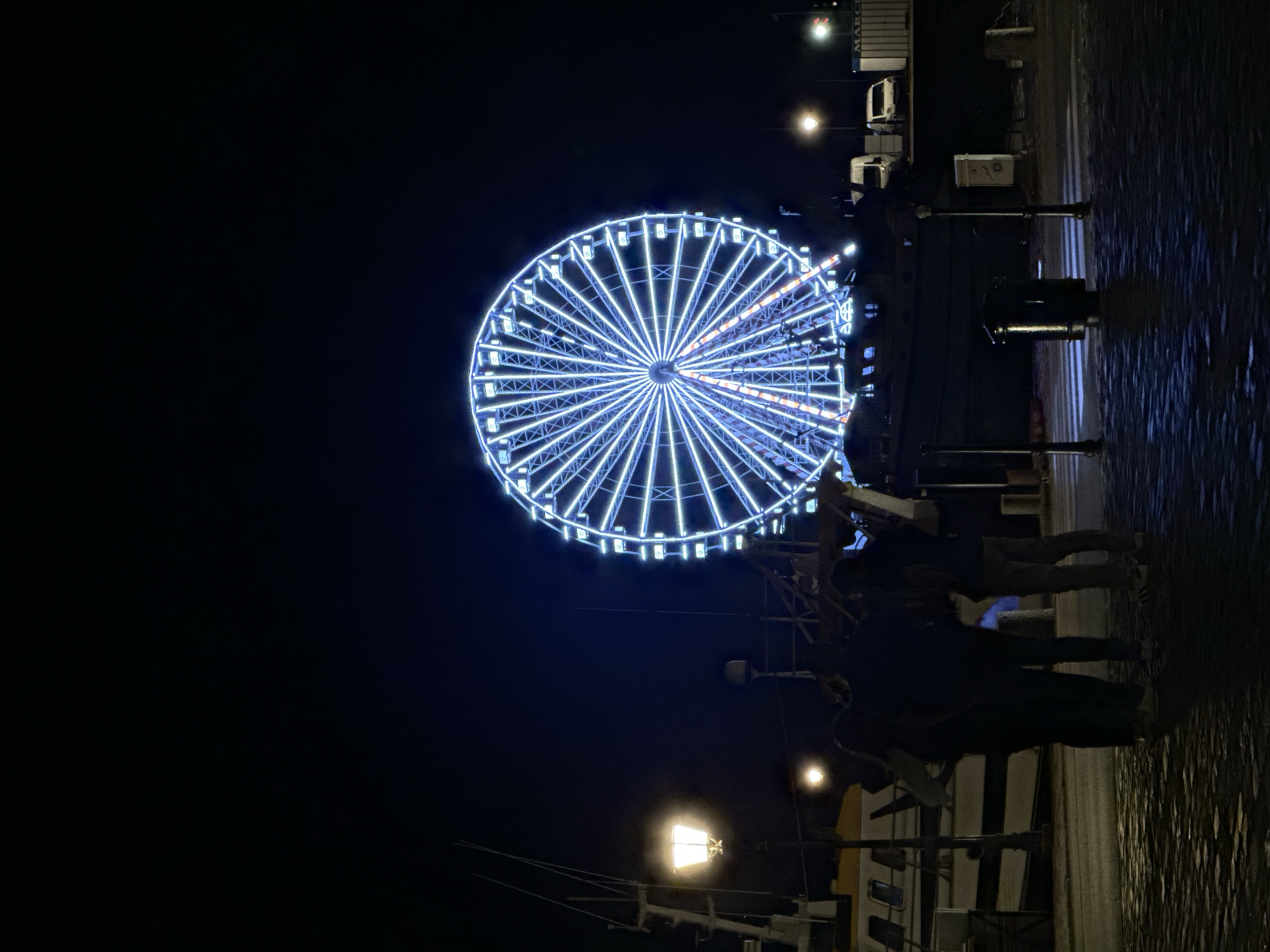 A nighttime scene in Honfleur, France, depicts a brightly lit Ferris wheel dominating the background. The Ferris wheel is illuminated with cool blue and white lights, with some red accents visible near its center. In the foreground, three individuals are standing close together, their backs to the camera, appearing to observe the Ferris wheel. They are silhouetted against the dimly lit surroundings. To the left, a lamp post casts a warm yellow glow. A boat is docked to the left of the frame. The ground is paved with cobblestones, and a trash receptacle is visible. In the mid-ground, there are vehicles and shipping containers. The sky is dark and clear, indicating a night time setting.