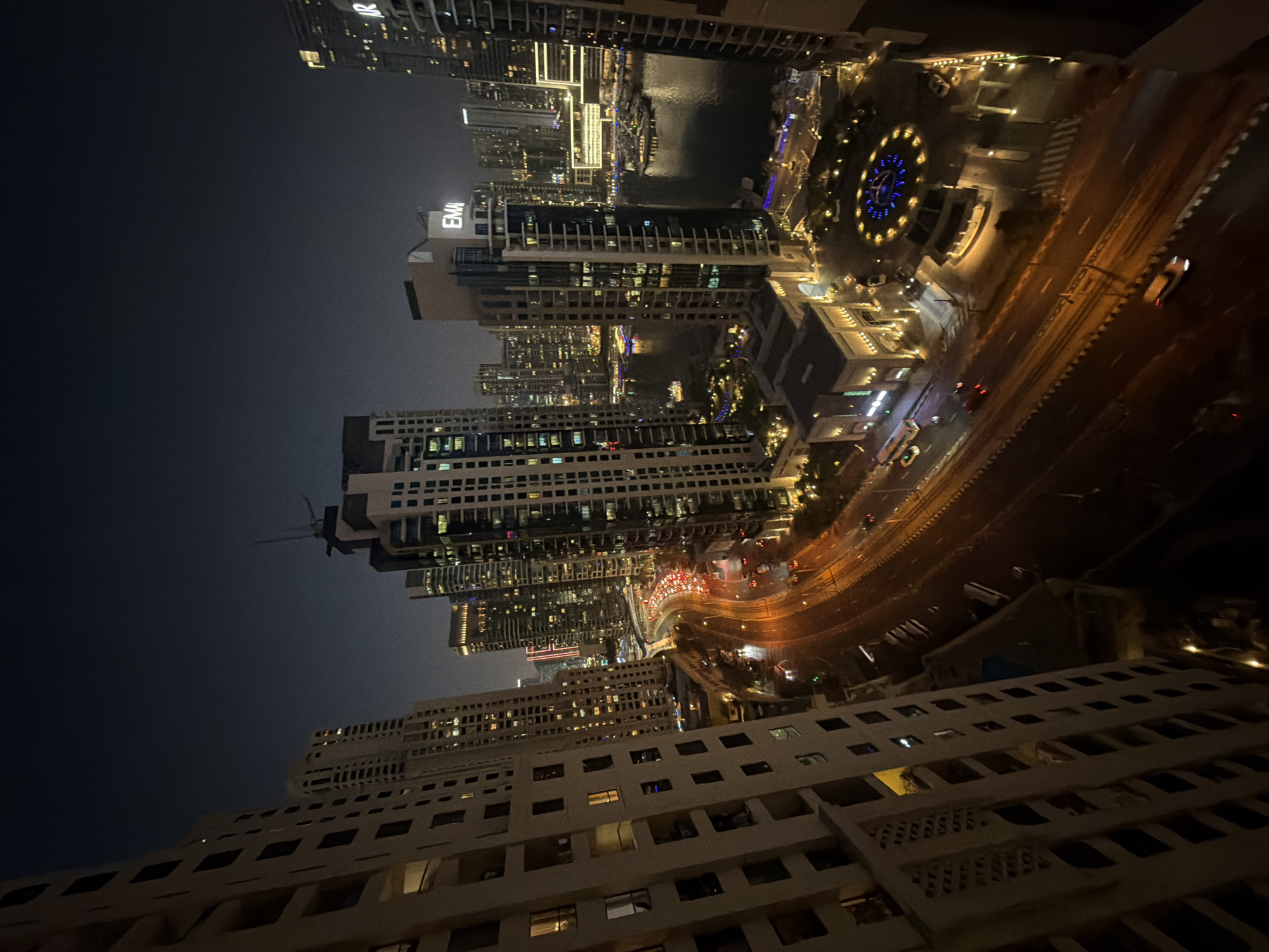 The aerial view captures a vibrant nighttime scene in Dubai Marina, United Arab Emirates. A multi-lane highway, illuminated by streetlights and the taillights of traffic, curves through the urban landscape. On either side of the highway, towering skyscrapers with their windows aglow pierce the dark sky. One prominent building displays the "EMAAR" logo. In the foreground, the textured facade of a residential building is visible. The scene extends to a marina filled with boats and yachts, reflecting the city lights on the calm water. The overall impression is one of a bustling, modern metropolis at night.