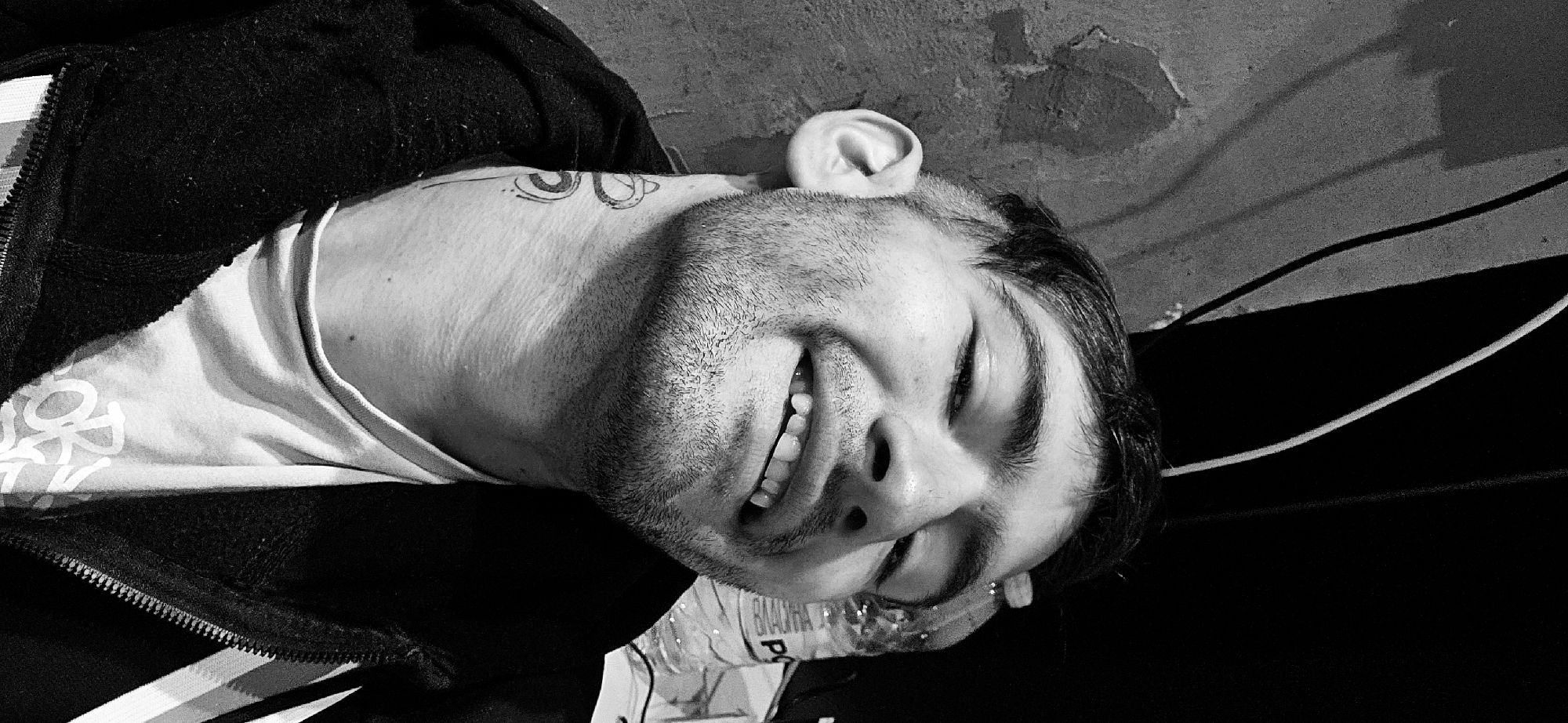 A black and white photograph displays a man lying down, appearing to be on his back or side, with his head tilted. He is smiling broadly, revealing his teeth, and his eyes are squinted, conveying a sense of amusement or happiness. He has short beard stubble and dark hair. He is dressed in a light-colored (possibly white or off-white) t-shirt beneath a dark, likely black, zipped jacket. A distinct tattoo featuring abstract, intertwined shapes is visible on his neck or shoulder. His right ear is prominent and appears slightly pressed against the surface he is resting on.
In the background, a textured wall, possibly concrete or brick, shows some wear. Dark and light colored wires or cables run across the background. A partially visible clear plastic bottle is positioned near his chin, bearing Cyrillic text "ВЛАДХА" and a "P" (likely from a brand name). The overall scene suggests an informal indoor setting in Plovdiv, Bulgaria. The black and white tonality adds a stylistic or candid feel to the image.