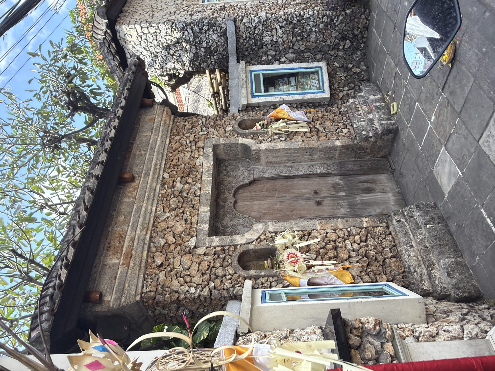 The image depicts a stone temple entrance in Kuta, Indonesia. The temple features a rustic stone facade with a traditional tiled roof. Elaborate wooden doors are set into the main entrance, flanked by decorative niches containing offerings. Small windows with blue frames are visible on either side of the entrance. The ground is paved with stone tiles, and a few offerings are scattered on the pavement. In the lower right corner, a motorcycle mirror reflects a glimpse of the street and the temple's surroundings. Trees with lush green foliage grow on top of the temple roof and nearby structures. The scene is set during the day, with sunlight casting shadows and illuminating the stone work.