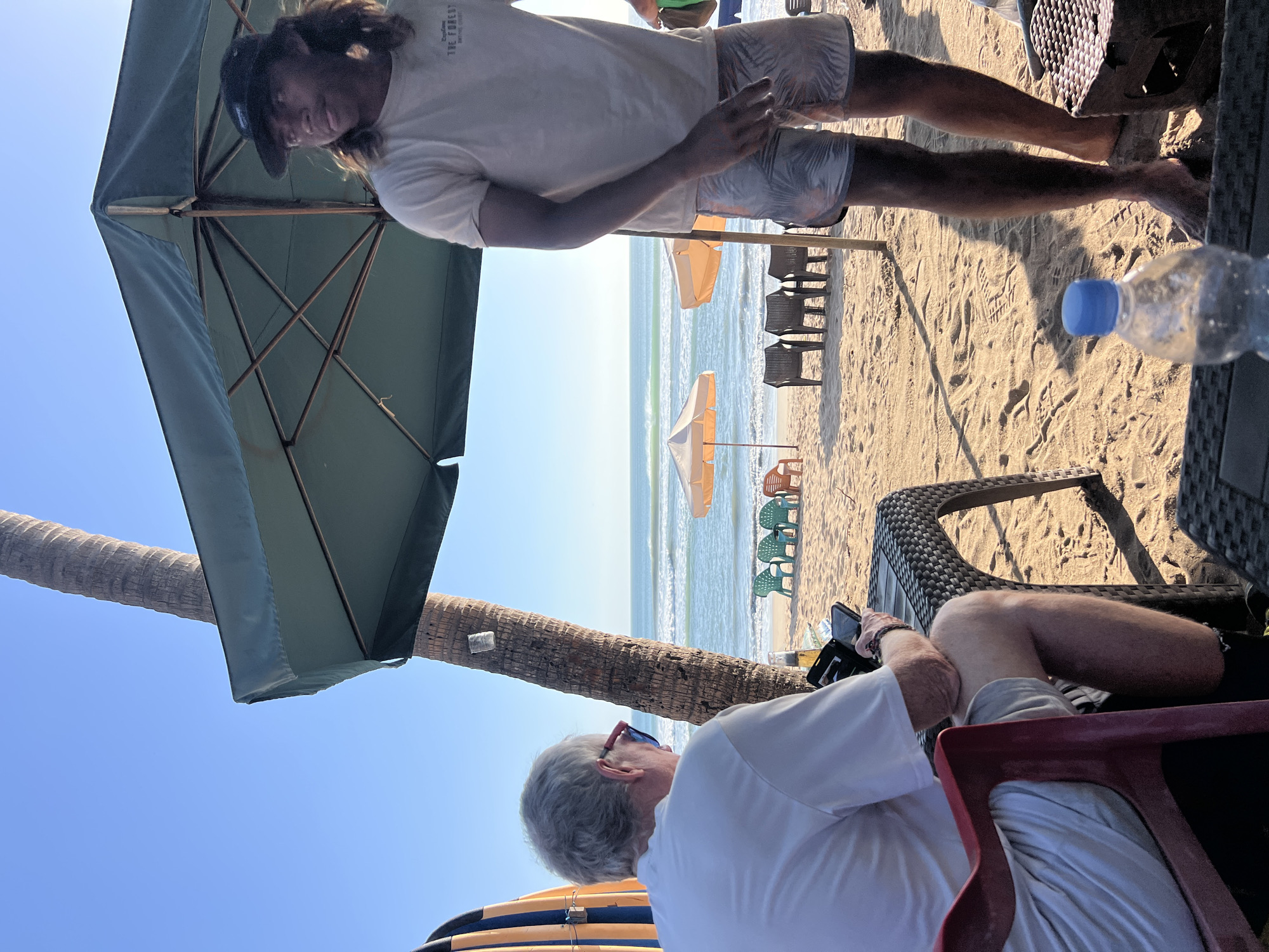 In Kuta, Indonesia, two men are seated on a beach with a backdrop of the ocean and sky. The man on the left, facing away from the camera, has grey hair and is wearing a white polo shirt and grey shorts. He is seated at a woven table, holding a phone in his right hand and a wallet in his left. A surfboard, partially visible, stands to his left. The man on the right, facing the camera, has long, dark hair and is wearing a white t-shirt and patterned shorts. He stands under a large green umbrella, looking towards the ocean. A bottle of water sits on a table in the foreground. In the background, beach chairs and umbrellas are arranged on the sand, with gentle waves lapping the shore under a clear blue sky.