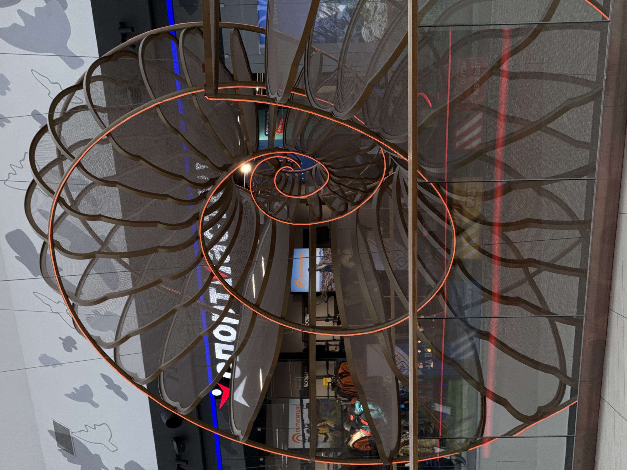 The image shows a spiral sculpture, likely a nautilus shell, within a building, possibly a shopping mall or public space, in Perm, Russia. The sculpture is constructed of a dark gray mesh material with a light brown frame and is outlined with bright orange neon lighting, emphasizing its spiral form. Through the mesh, reflections and signs of the mall are visible, including storefronts with the words "ПОХОДЫ" (meaning "hikes" or "expeditions" in Russian) and other logos. The background features a wall with a light-colored design that has gray silhouettes of animals, notably birds and a possible beaver. The overall scene is indoors and well-lit, suggesting it is a display or architectural feature within a commercial environment.