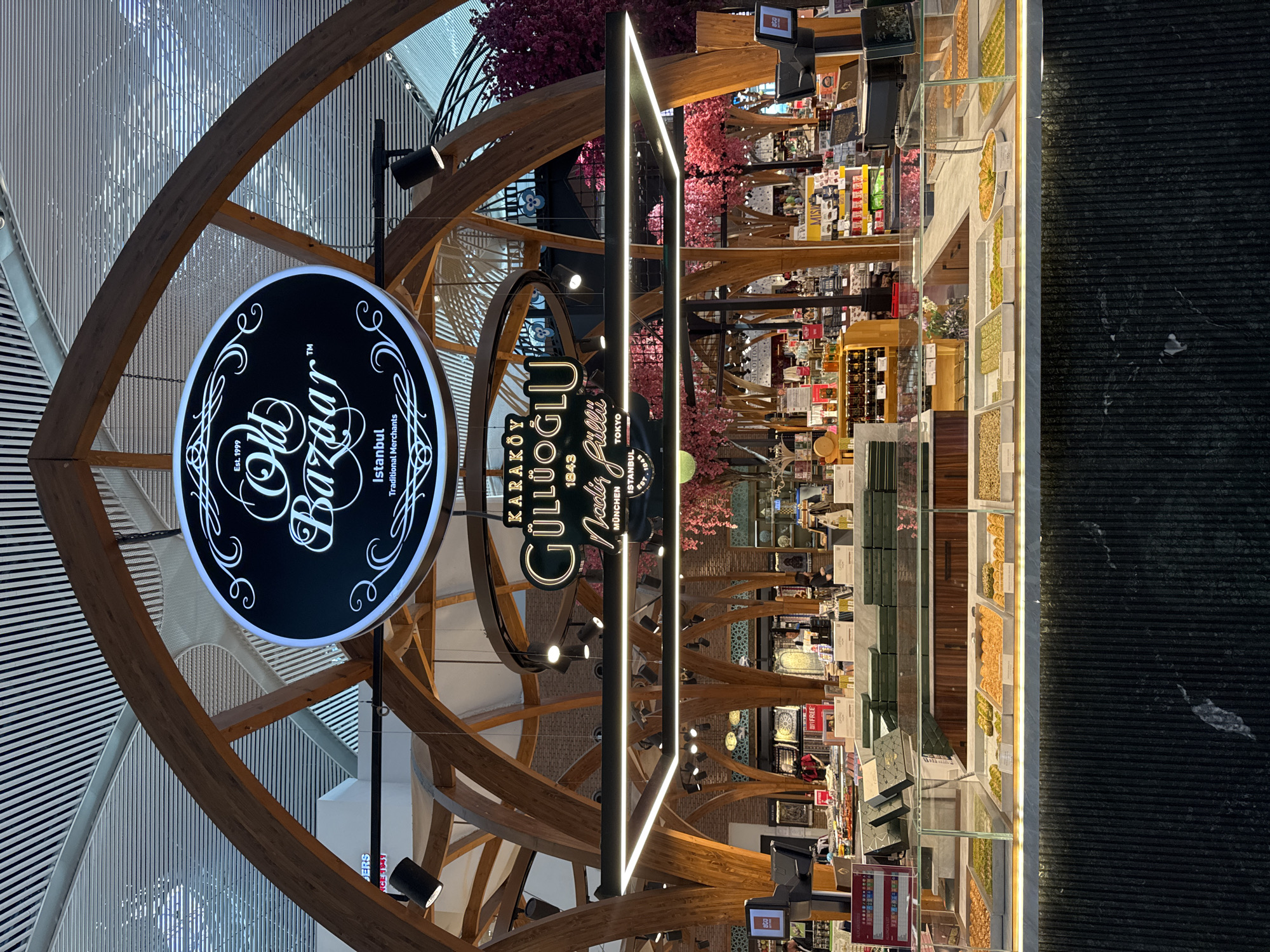 This image depicts an inviting indoor retail space, likely a high-end shop specializing in Turkish sweets and traditional goods, possibly located within an airport or large shopping complex. The scene is dominated by a modern architectural design featuring elegant wooden arches that frame the shop.
Two prominent signs identify the businesses. On the left, a circular black sign with white script reads "Old Bazaar™ Istanbul Traditional Merchants Est. 1999". To its right, a tall, vertical sign features "KARAKÖY GÜLLÜOĞLU" with "Nadir Güllü" beneath it, and indicates "1843" (likely establishment year), along with locations "ISTANBUL", "TOKYO", and "MÜNCHEN".
The shop's interior is well-lit and features extensive display cases filled with a variety of confectionery, predominantly baklava and other nut-based pastries, arranged in neat rows. Some items appear to be pistachios or similar nuts. Behind the main display counters, shelves hold packaged goods and other products. The decor includes artificial pink cherry blossom trees, adding a touch of color and natural aesthetic to the modern setting. Sleek, linear LED lights illuminate the central display area, complementing various spotlights mounted on the wooden structures. The floor in the foreground is a dark, textured material.
The overall impression is one of quality and tradition meeting contemporary design, inviting customers to explore and purchase. The visible text clearly indicates a Turkish origin, with Karaköy Güllüoğlu being a renowned name in baklava, suggesting this is a branch of their famous establishment. The mention of international cities on the Güllüoğlu sign highlights the brand's global presence.