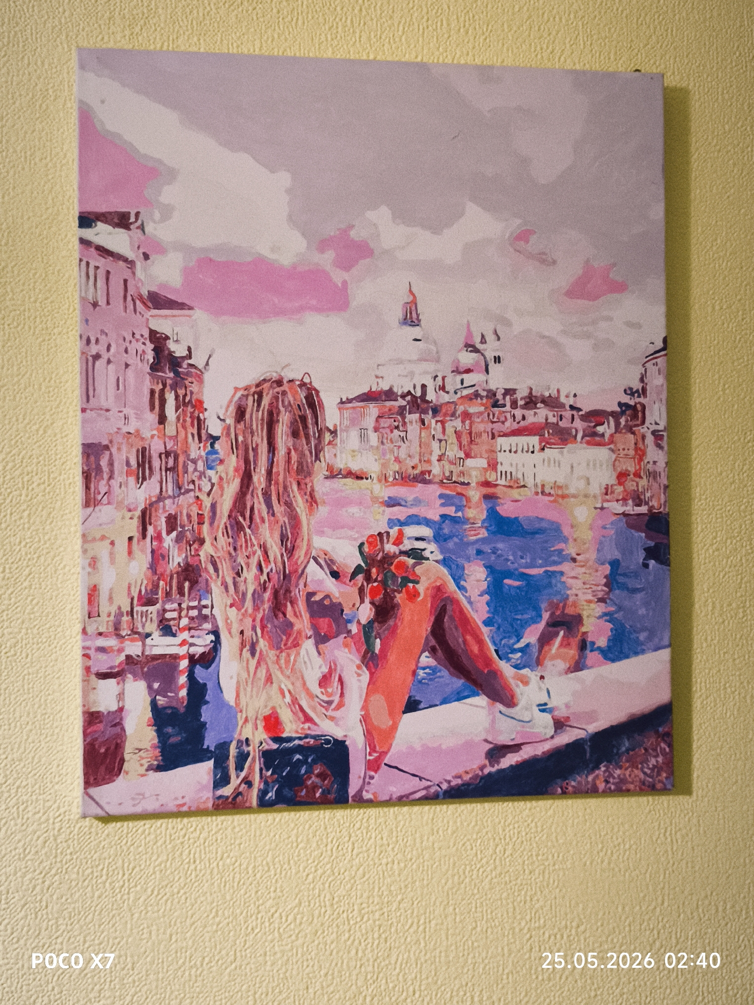 A square painting hangs on a yellow wall. The painting depicts a girl with long blonde hair sitting on a ledge. She is holding a bouquet of red flowers. In the background is a cityscape with a canal, buildings, and a dome. The painting uses a palette of pink, purple, blue, and red hues. The city depicted is likely Venice, Italy, evidenced by the canals, architectural style, and domed building resembling Saint Mark's Basilica.