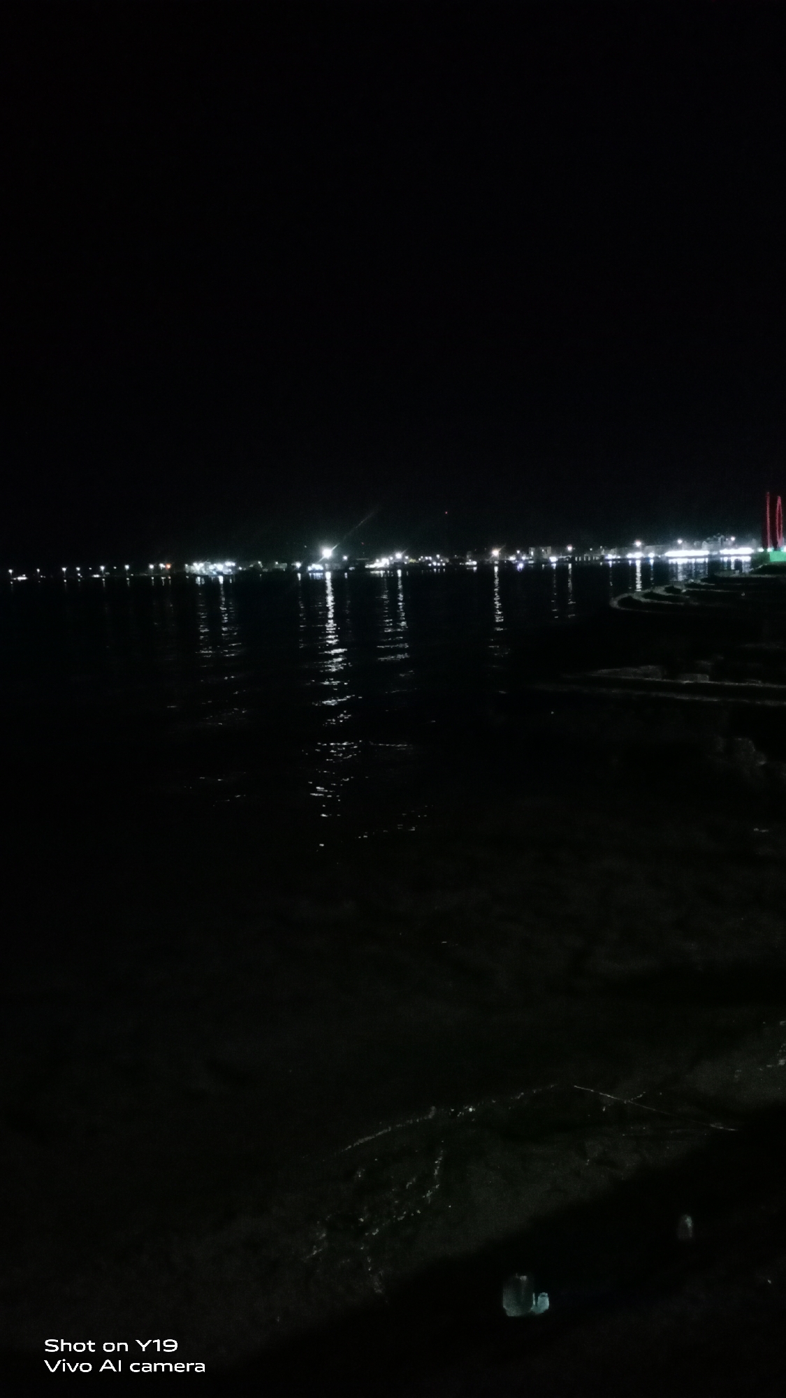 This is a nighttime, vertical, low-angle, medium shot of a body of water, likely the sea, in Sea, Indonesia. The dark water reflects the lights of a distant city skyline, which is visible along the horizon. The city lights appear as bright white dots and streaks, creating shimmering reflections on the water's surface. The foreground shows the dark, rippling water with some subtle, illuminated ripples near the bottom right. The overall impression is one of a tranquil, urban waterfront at night. The bottom left corner of the image contains text that reads "Shot on Y19 Vivo Al camera".
