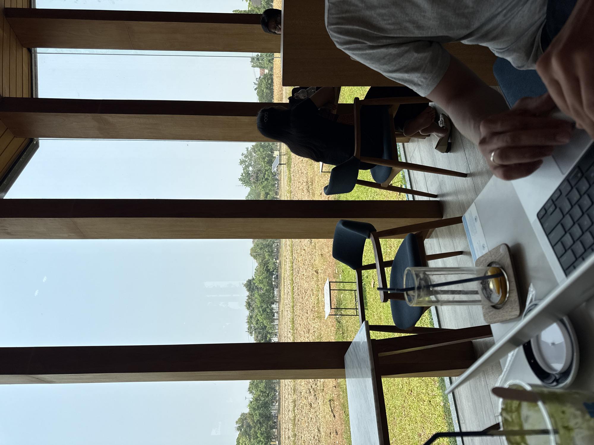 The image, taken in Chiang Mai City Municipality, Thailand, shows an interior scene with a view of a rural landscape through large windows. The windows have horizontal wooden beams separating the panes.
Inside, a table occupies the foreground, on which sit a laptop, a glass with straws, and a drink. A person's hands are visible on the laptop. Another person in a grey t-shirt is partially visible in the top right corner, working on a laptop.
Two dark-colored chairs are positioned near the table. Through the window, a field with trees and what appears to be agricultural land can be seen. A person's head peeks through the top of a window. A second person is sitting on a chair. A solar panel is visible in the background. The sky is overcast.