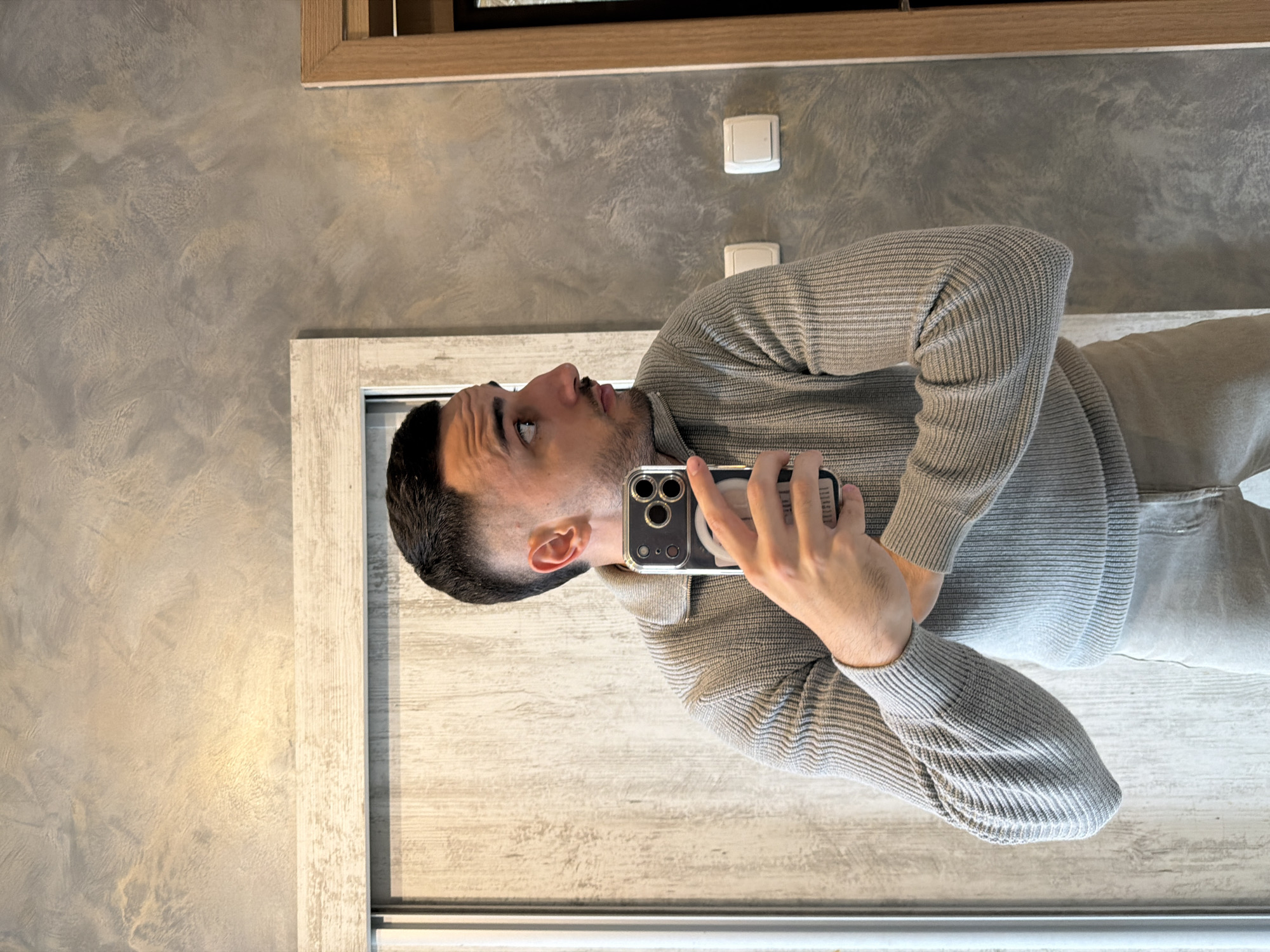 A man is standing in front of a mirror and taking a selfie. He is wearing a gray sweater and gray pants. The background is a gray wall with two light switches. The mirror has a light wood frame. The location in Plovdiv, Bulgaria is not discernible from the image.