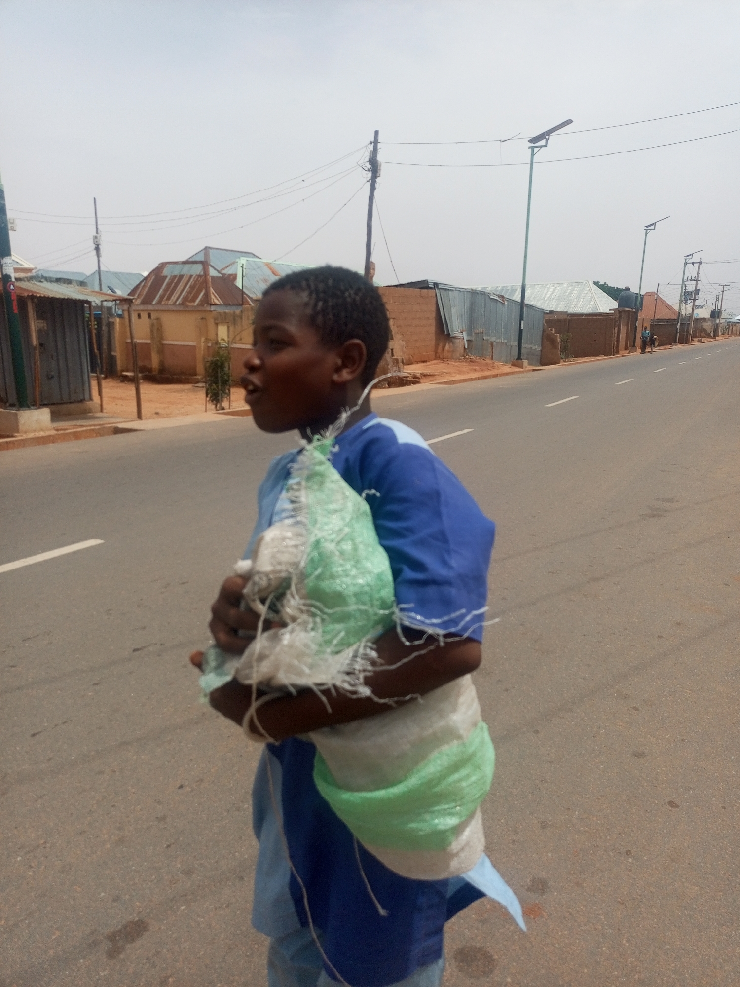 The image depicts a young person standing on the side of a street in what appears to be a residential area. The individual is carrying a bag, possibly containing discarded materials. The location appears to be in a city. The photograph captures the individual's appearance, clothing, and the bag they are carrying. The image appears to be taken in Tempure, Nigeria. There are powerlines and solar streetlamps visible in the background. Several buildings can be seen in the distance. Two people are walking in the distance.