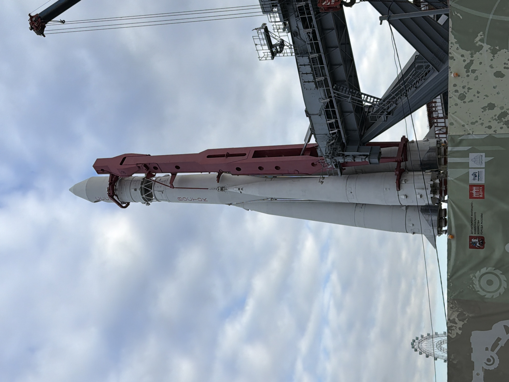 The image captures a rocket displayed at the VDNKh (Exhibition of Achievements of National Economy) in Moscow, Russia. The rocket, a Soyuz model, is white with black and red markings. It is positioned under a large metal structure, likely part of the exhibition or launch support equipment. A crane is visible in the upper left. The background consists of a cloudy sky. A partial view of a Ferris wheel and an informational stand can be seen in the lower right of the image.