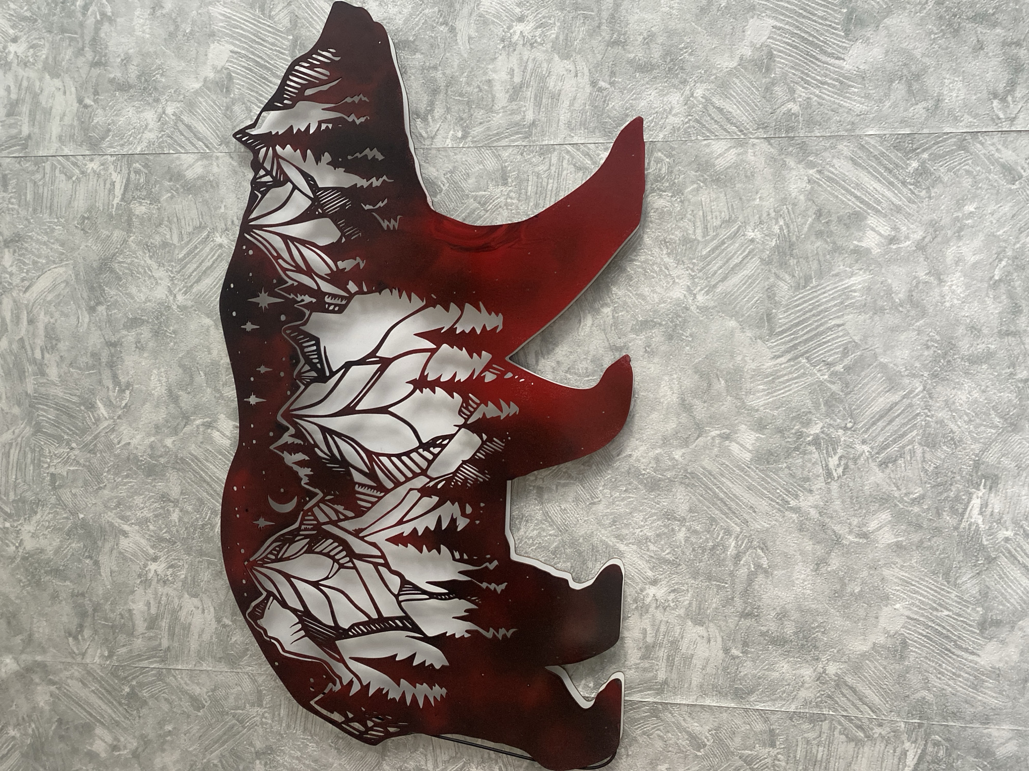 A metallic wall art piece shaped like a bear is mounted on a textured gray wall. The art piece features a silhouette of a bear filled with a landscape of mountains, pine trees, a crescent moon, and stars. The landscape is rendered in white and dark red, with the red color fading from a deep burgundy on the bear's exterior to a lighter red towards the center of the design. The wall behind the art has a rough, brushstroke-like texture in various shades of gray. The location in Mamontovo, Russia, is not discernible from the image.