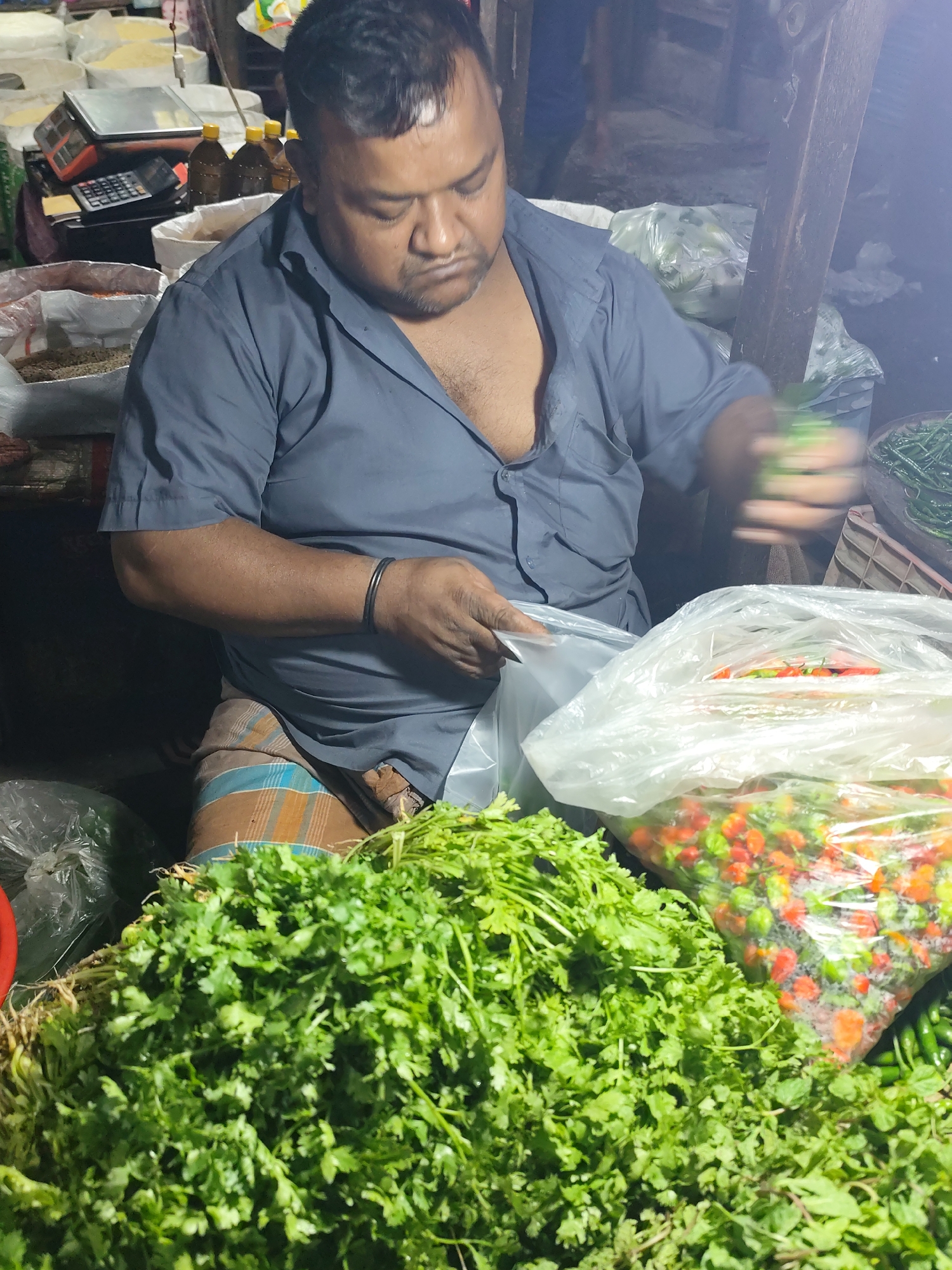 A vendor is shown at a market stall. He is an adult male with dark hair and a gray shirt, who is sitting with a small calculator and scale behind him. He is putting green beans into a clear plastic bag. In front of him is a large pile of cilantro and another clear plastic bag of chili peppers. The man is wearing a bangle on his left wrist and a checked lungi. The location is likely in the city of Dhaka, Bangladesh, given the person's attire and food for sale, and the style of scale and other equipment present.